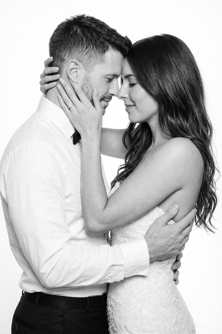 A black-and-white close-up of a romantic couple with their foreheads touching, eyes closed, and loving expressions. The woman is wearing a strapless lace dress and has long wavy hair, while the man is wearing a white shirt with rolled-up sleeves and 