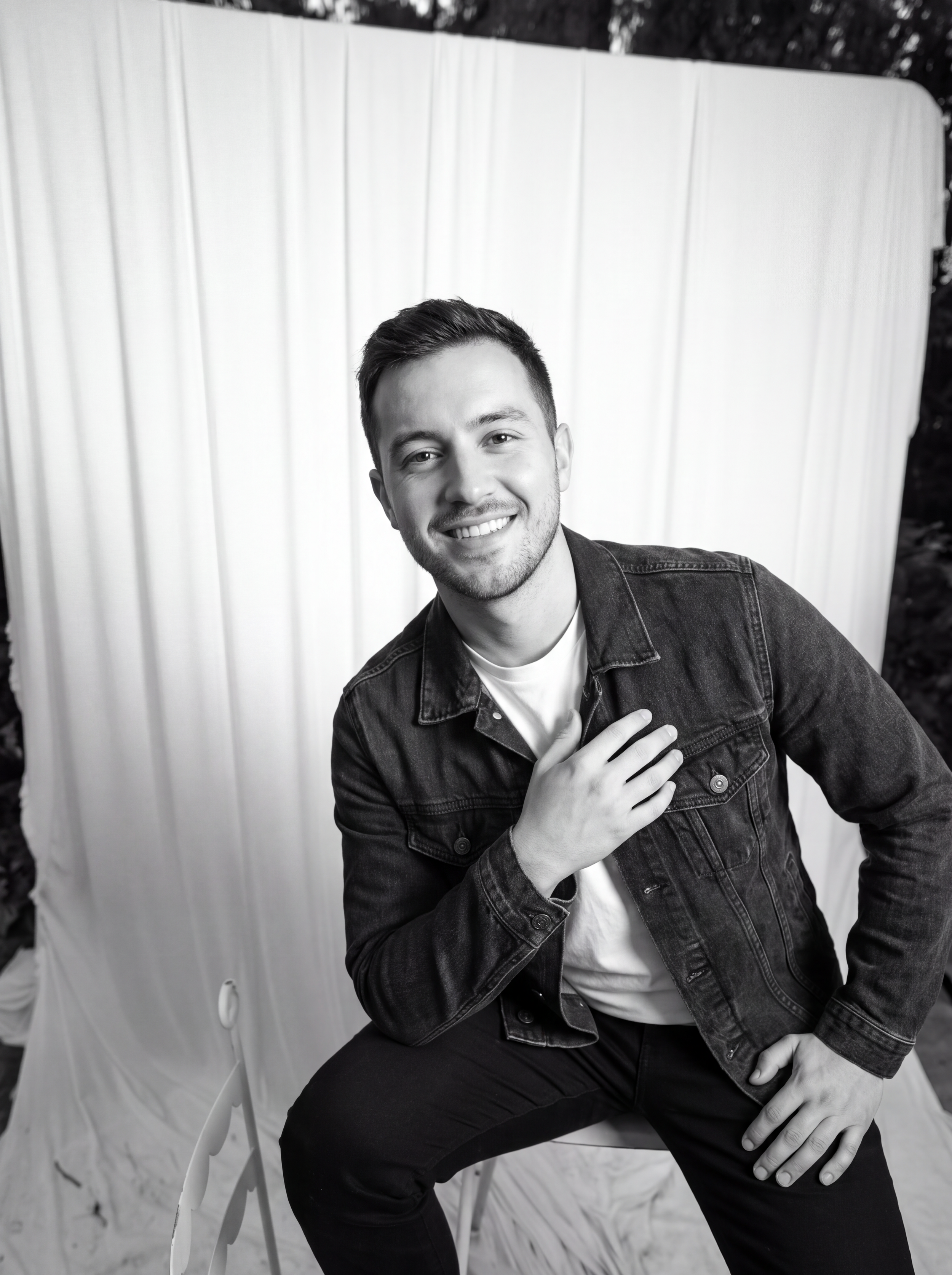 Black and white photo of a young man with short hair, smiling, wearing a denim jacket over a white shirt, sitting outdoors in front of a light-colored backdrop, with trees visible in the background.