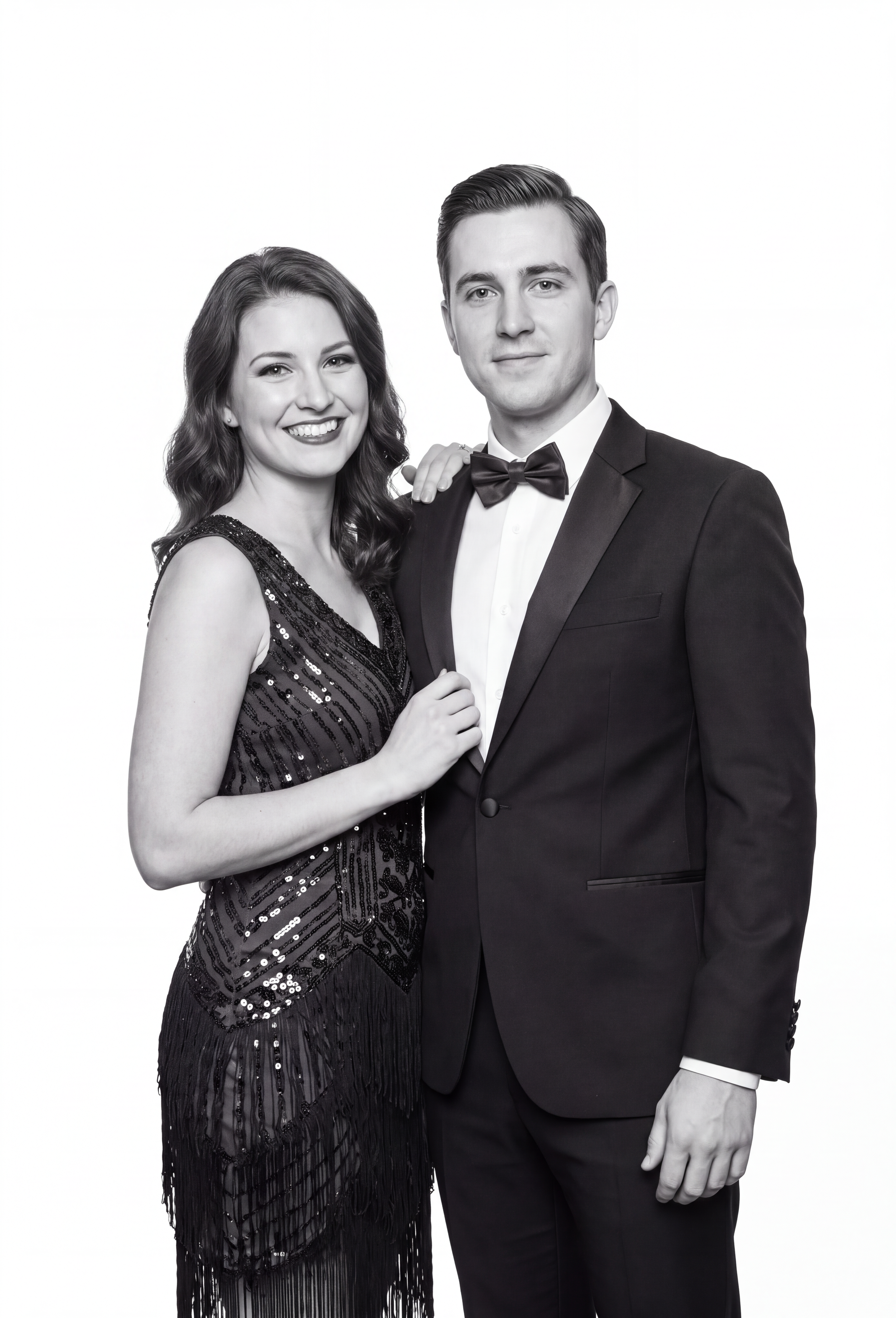 A black and white photo of a smiling woman in a sequined dress and a man in a tuxedo with a bow tie, standing close together against a plain white background.