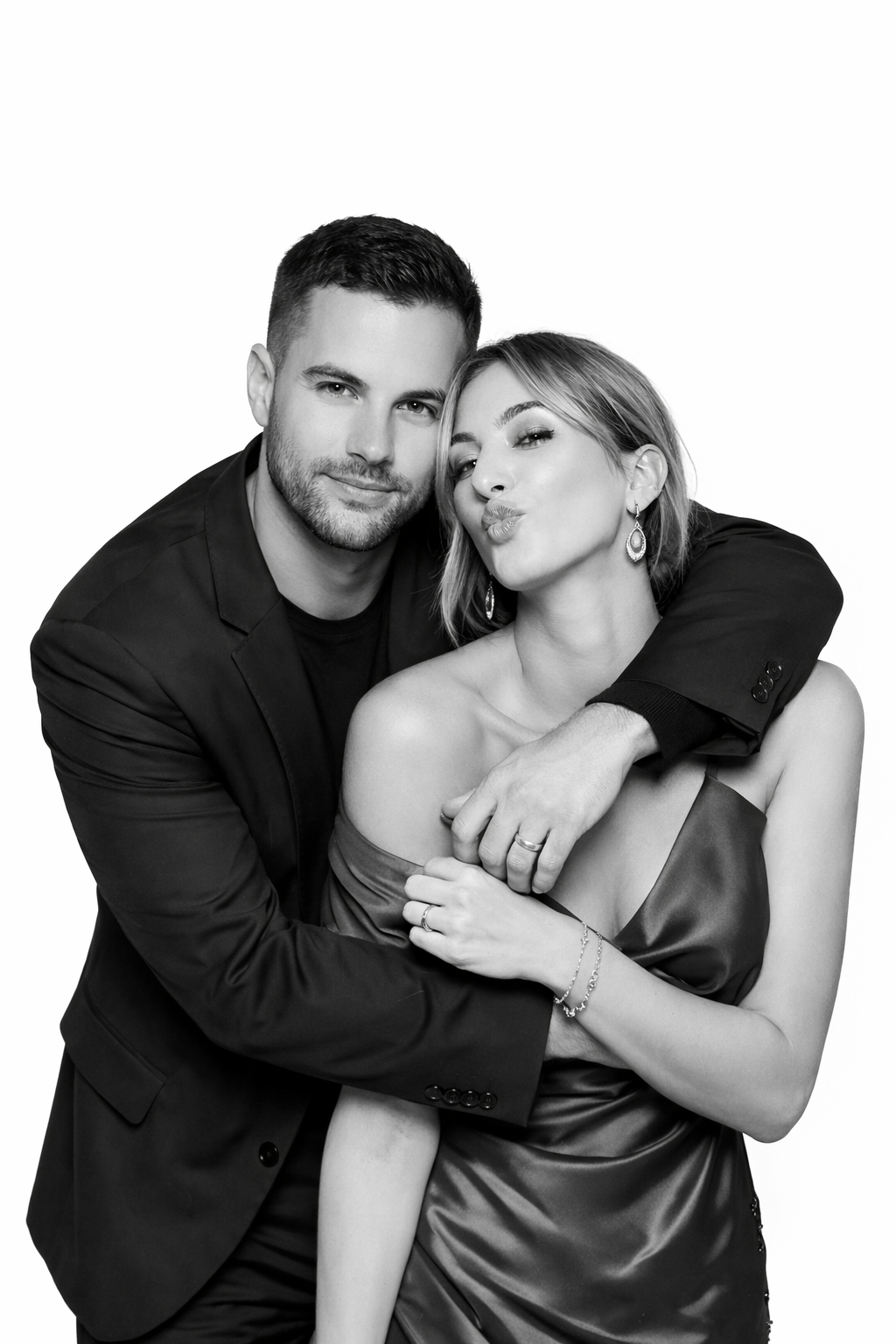 A black and white photo of a couple posing together, the man is wearing a blazer and has his arm around the woman's shoulders, who is making a kissy face. The woman is dressed in an off-the-shoulder satin dress, with jewelry and earrings.