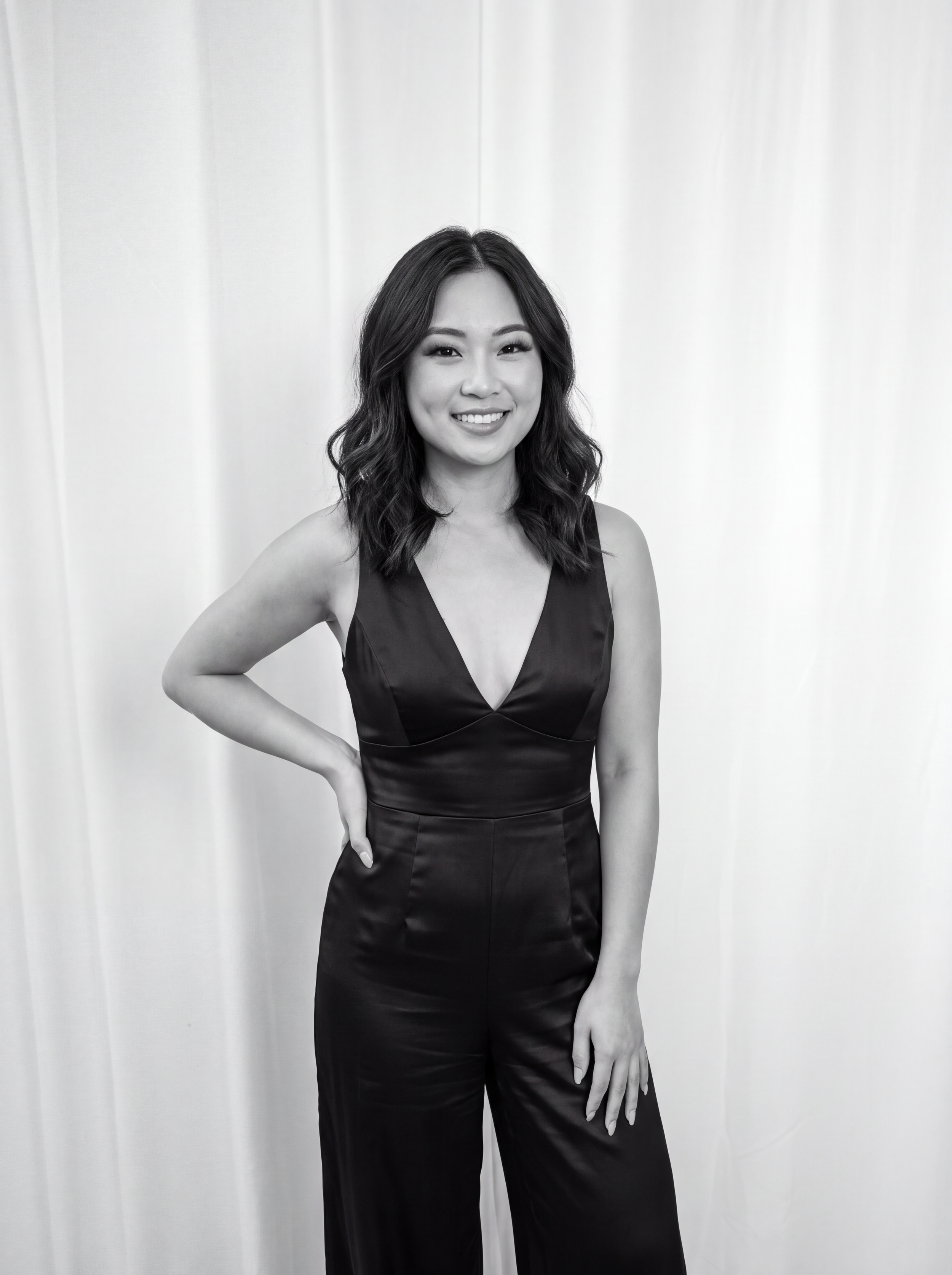 A woman with wavy hair smiling, wearing a sleeveless black dress, standing against a plain white background.