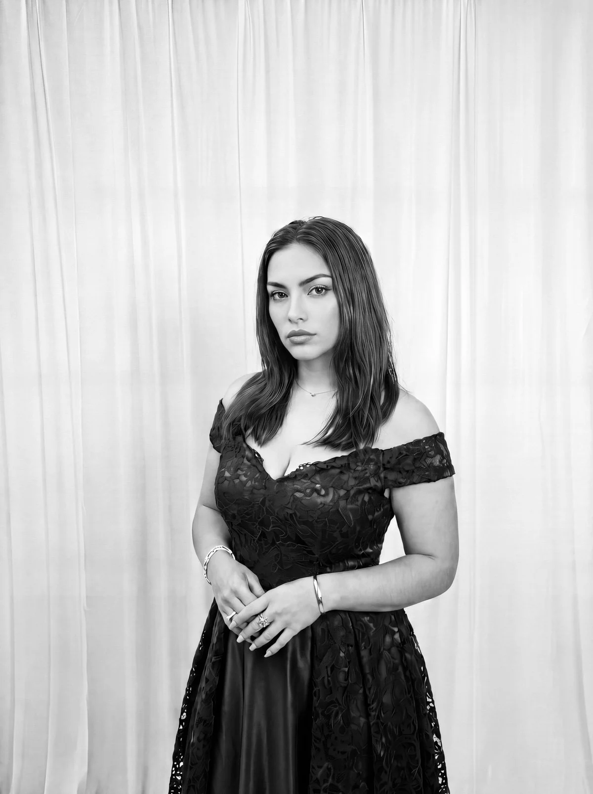 A woman in a black off-shoulder lace dress with wavy hair, standing in front of a curtain.