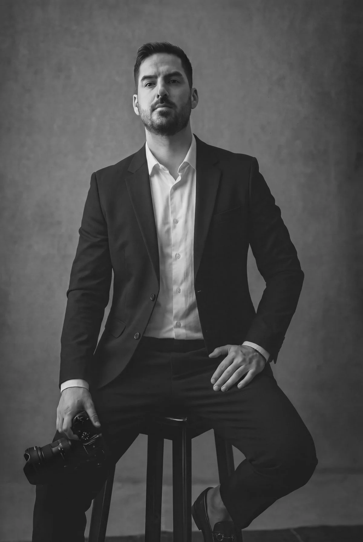 A man in a suit sitting on a stool, holding a camera, with a serious expression, in a black and white portrait.