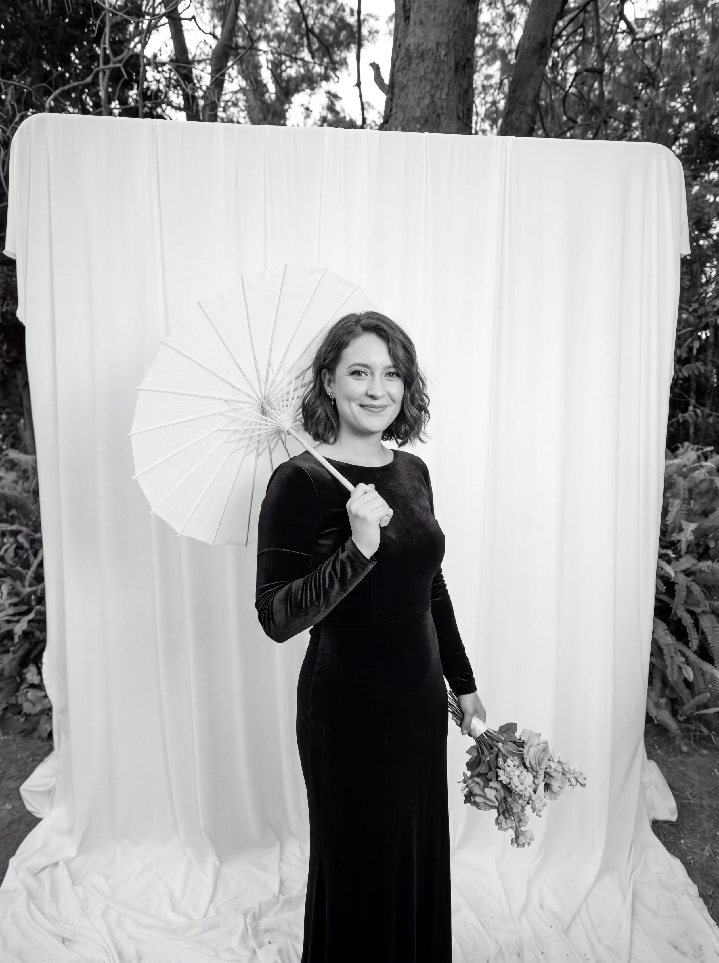 A woman in a black dress holding a bunch of flowers in one hand and a parasol in the other, standing in front of a white fabric backdrop outdoors with trees in the background.