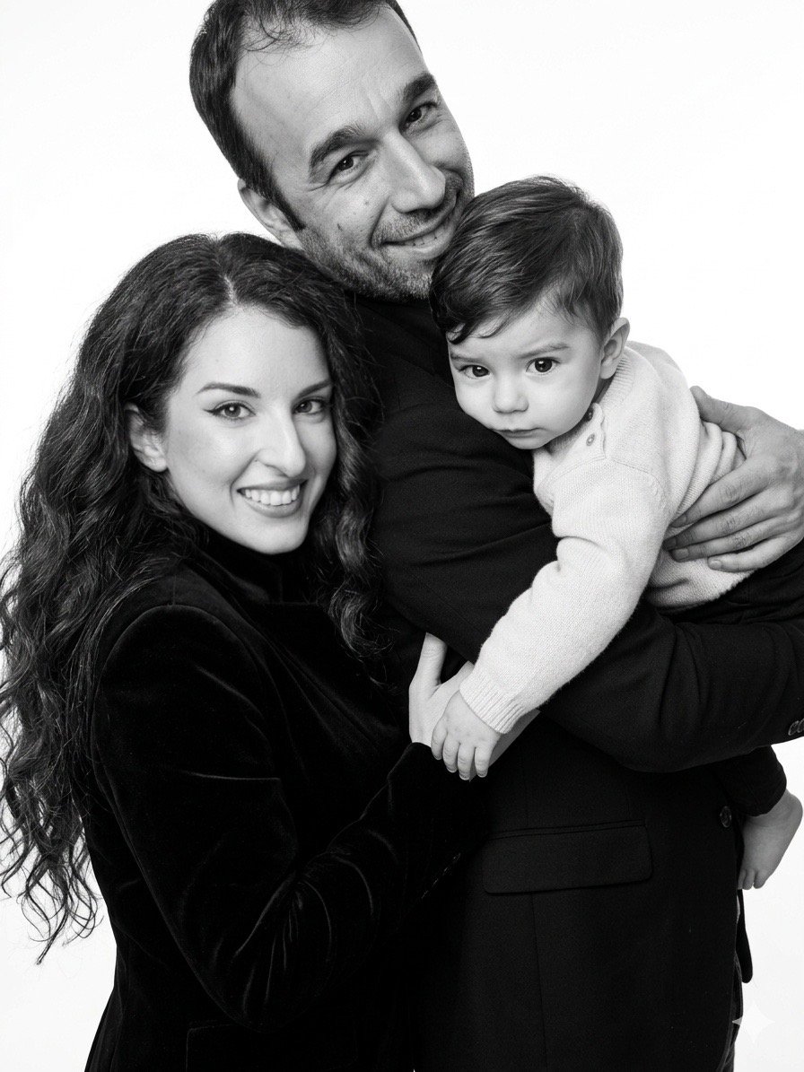 A black and white photo of a happy family of three: a woman with long wavy hair, a man with short hair and a beard, and a young boy. The woman is smiling, the man is smiling and holding the boy, who looks serious and is wearing a light-colored sweater.