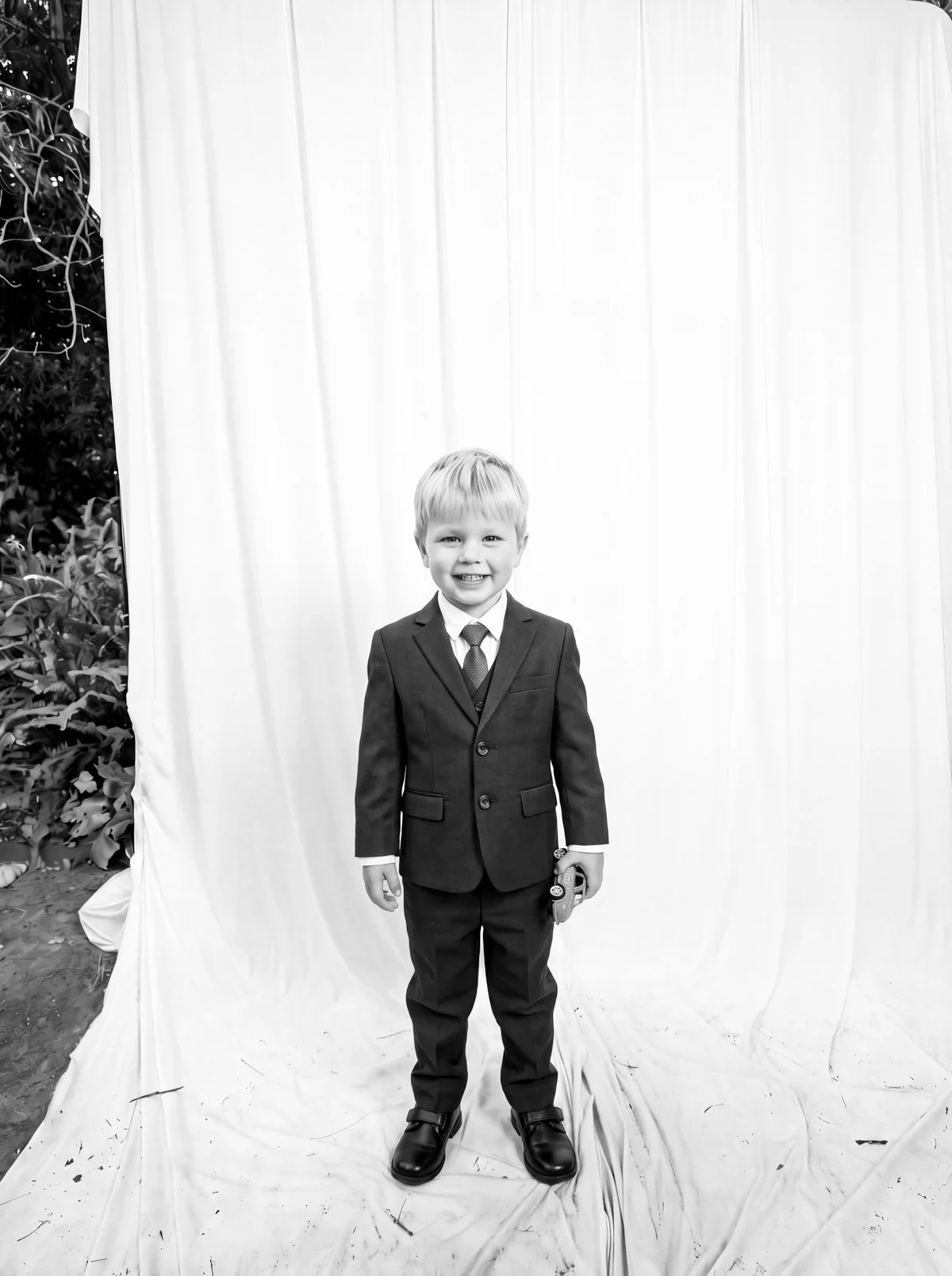 A young boy in a suit and tie standing in front of a plain curtain backdrop, smiling at the camera, outdoors with plants visible on the left side.