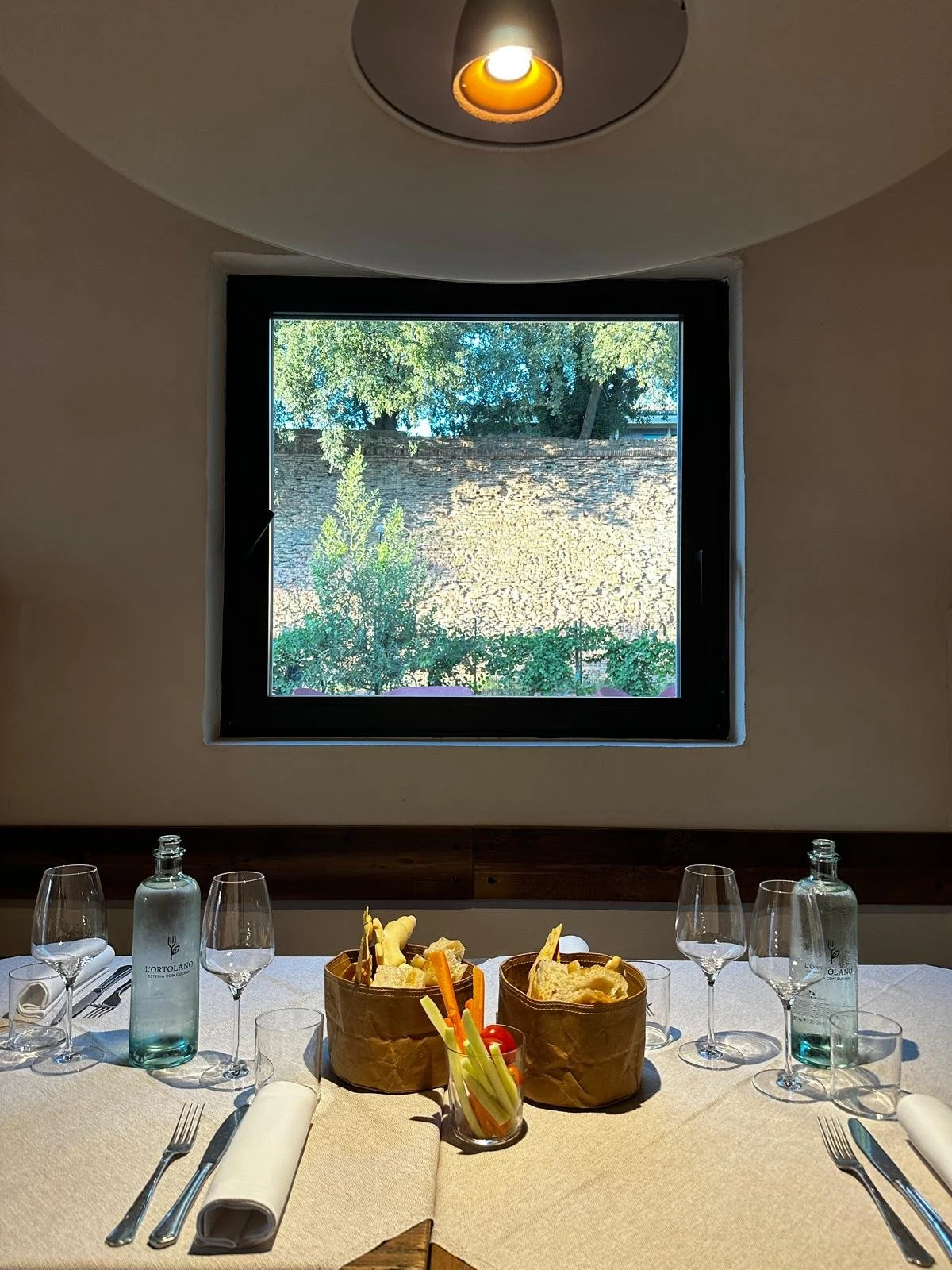 Tavola apparecchiata alla Cucina dellìOrtolano con bicchieri, caraffe d'acqua, piatti di pane e verdure, posate e tovaglioli, davanti a una finestra che si apre su un cortile con piante e le mura di Ferrara.
