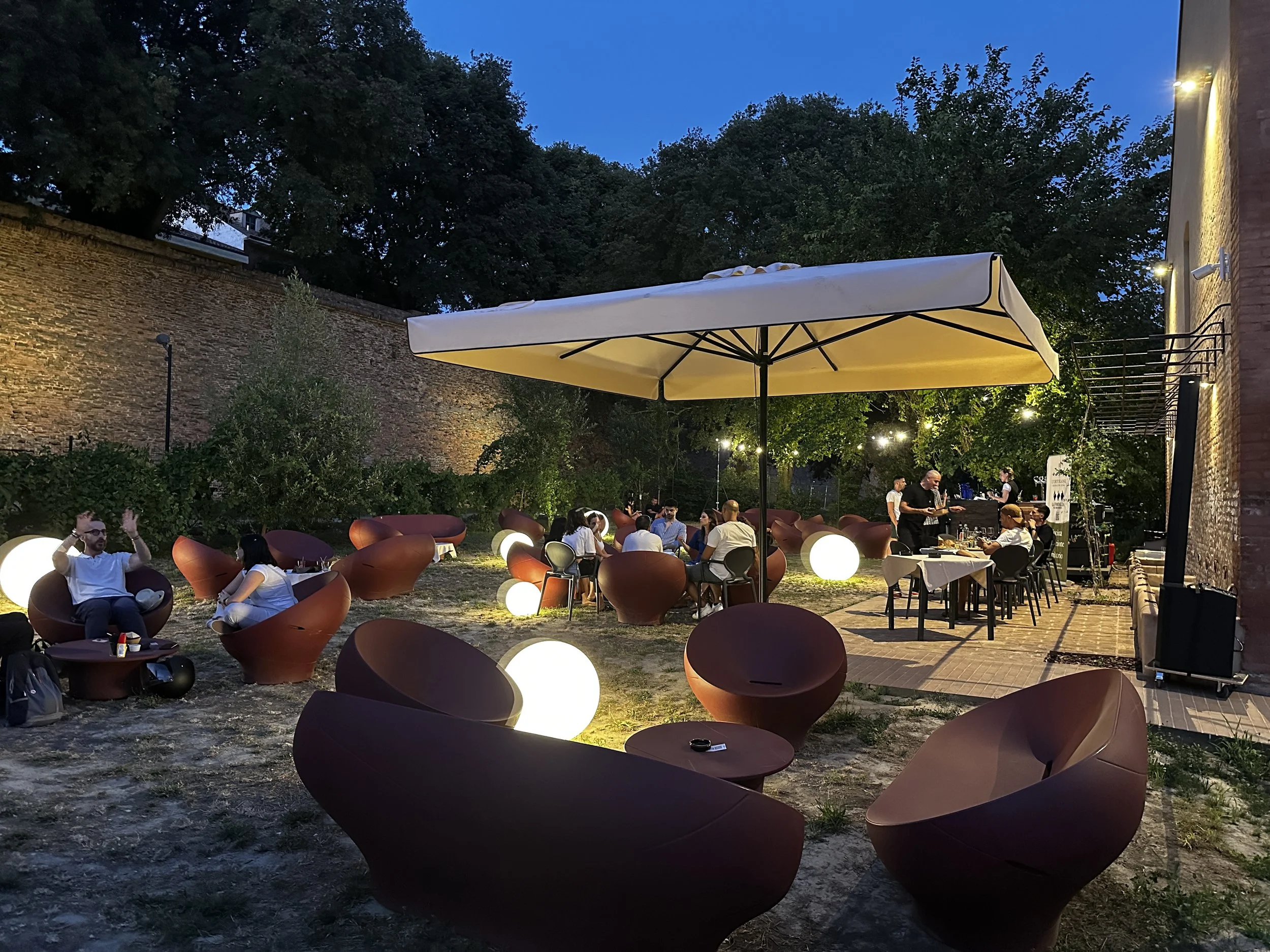 Bar dell'Ortolano in stile moderno e luci a sfera illuminate, persone sedute che chiacchierano e un ombrellone bianco. Atmosfera serale con luci soffuse e alberi sullo sfondo.