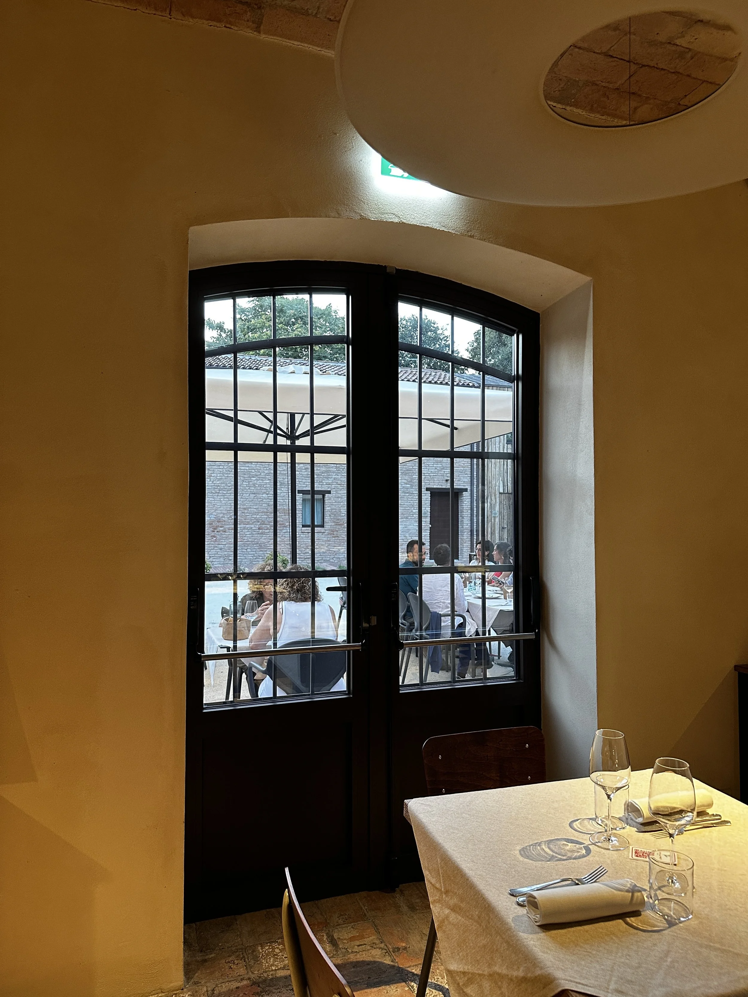 Vista di una sala da pranzo Cucina dell'Ortolano e attraverso una porta vetrata con inferriate e tavoli apparecchiati con tovaglie beige, bicchieri di vino, piatti e posate.
