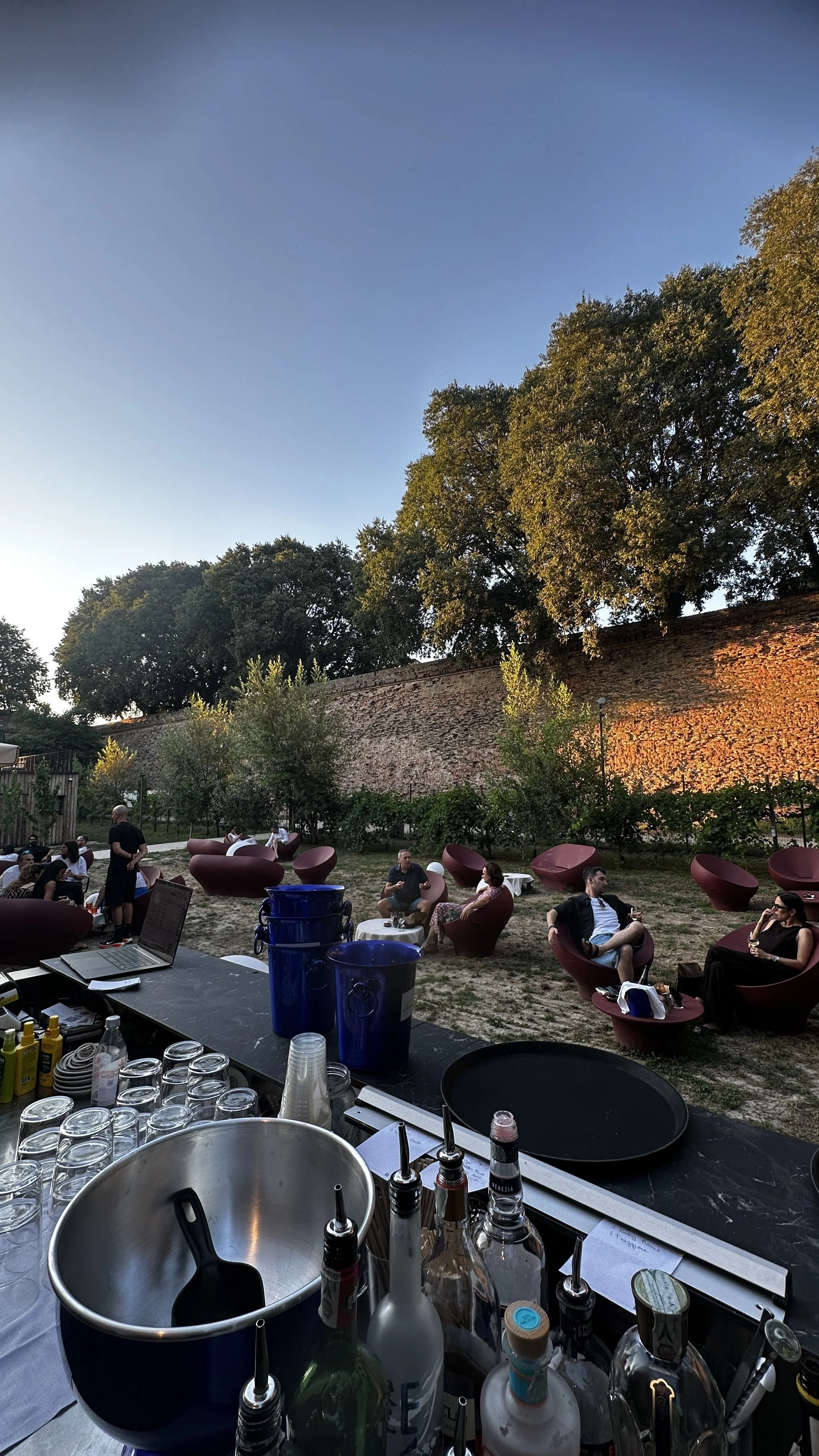 Vista bar dell'Ortolano con persone sedute all'aperto, alcune che si godono il tramonto e un muro di mattoni sullo sfondo, con alberi sopra.