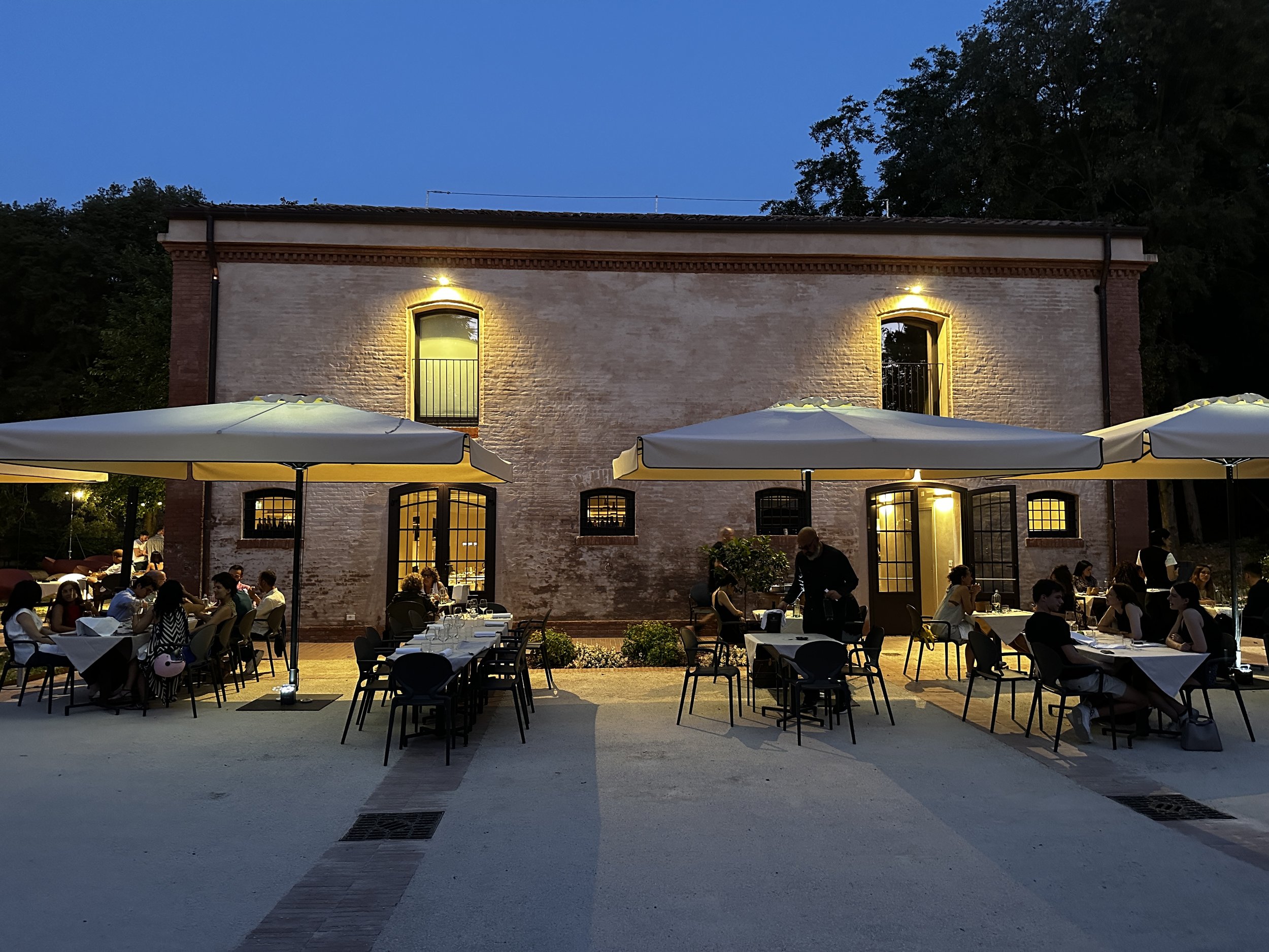 Cucina dell'Ortolano distesa all'aperto con tavoli e sedie sotto ombrelloni, persone che cenano e chiacchierano, un edificio in mattoni con luci calde e grandi finestre, e una sera con cielo blu e alcune luci luminose.