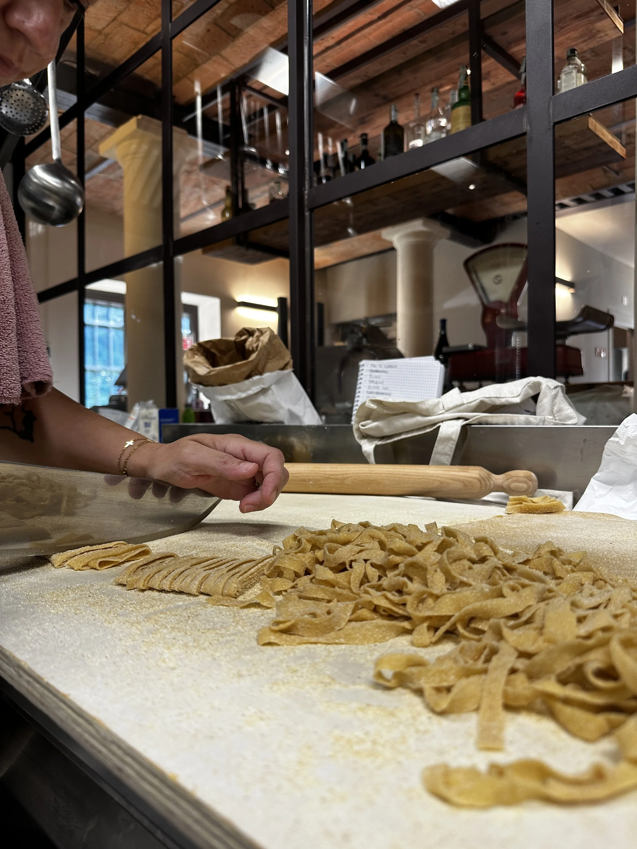 Cucina dell'Ortolano la nostra pasta fatta a mano sulla tavola, con varie forme di pasta. Sullo sfondo, un ambiente rustico con scaffali di bottiglie e pentole.