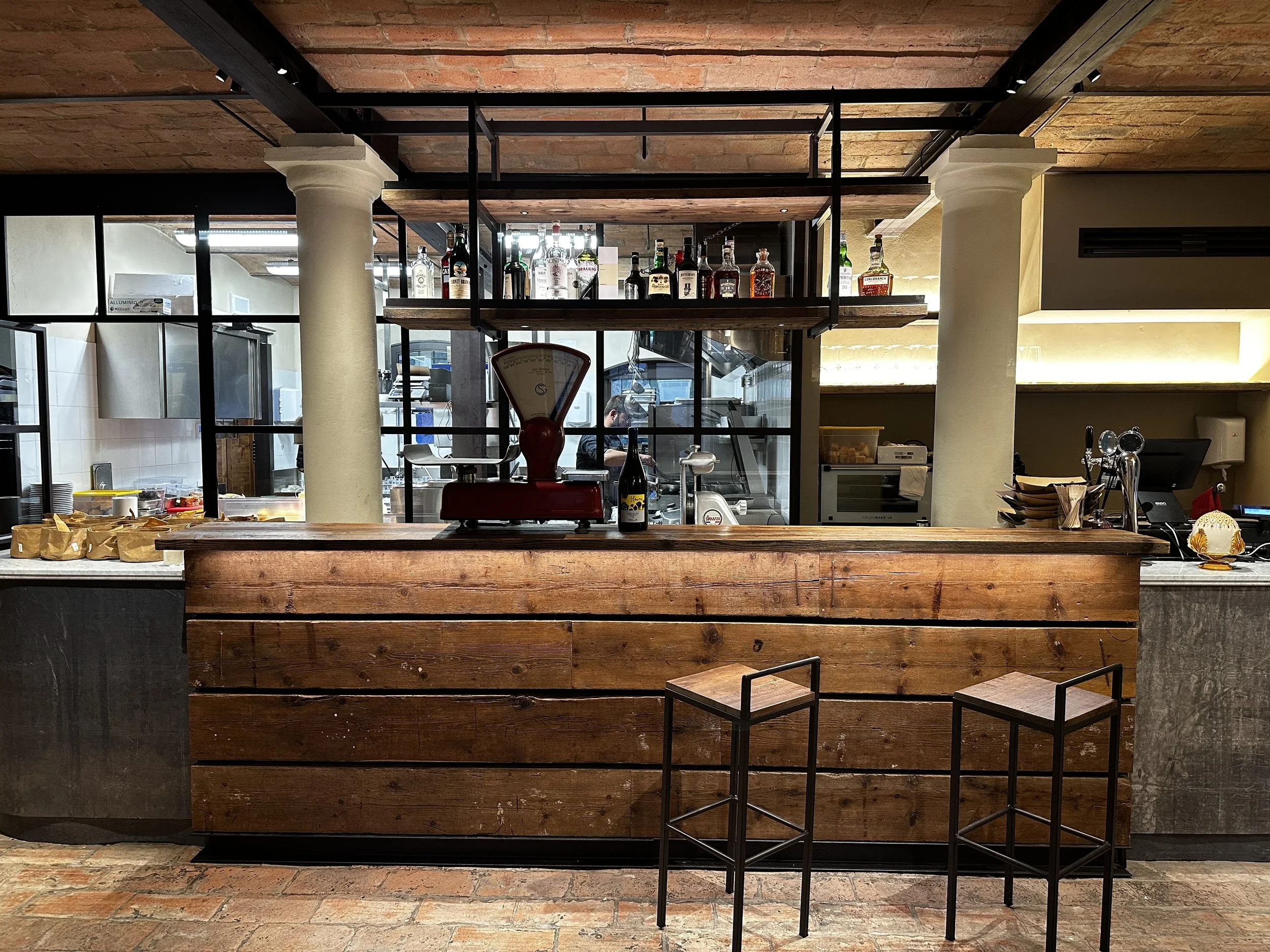 Salumeria Cucina dell'Ortolano con bancone in legno, scaffale con bottiglie di liquore e tavoli con sedie di ferro e legno.