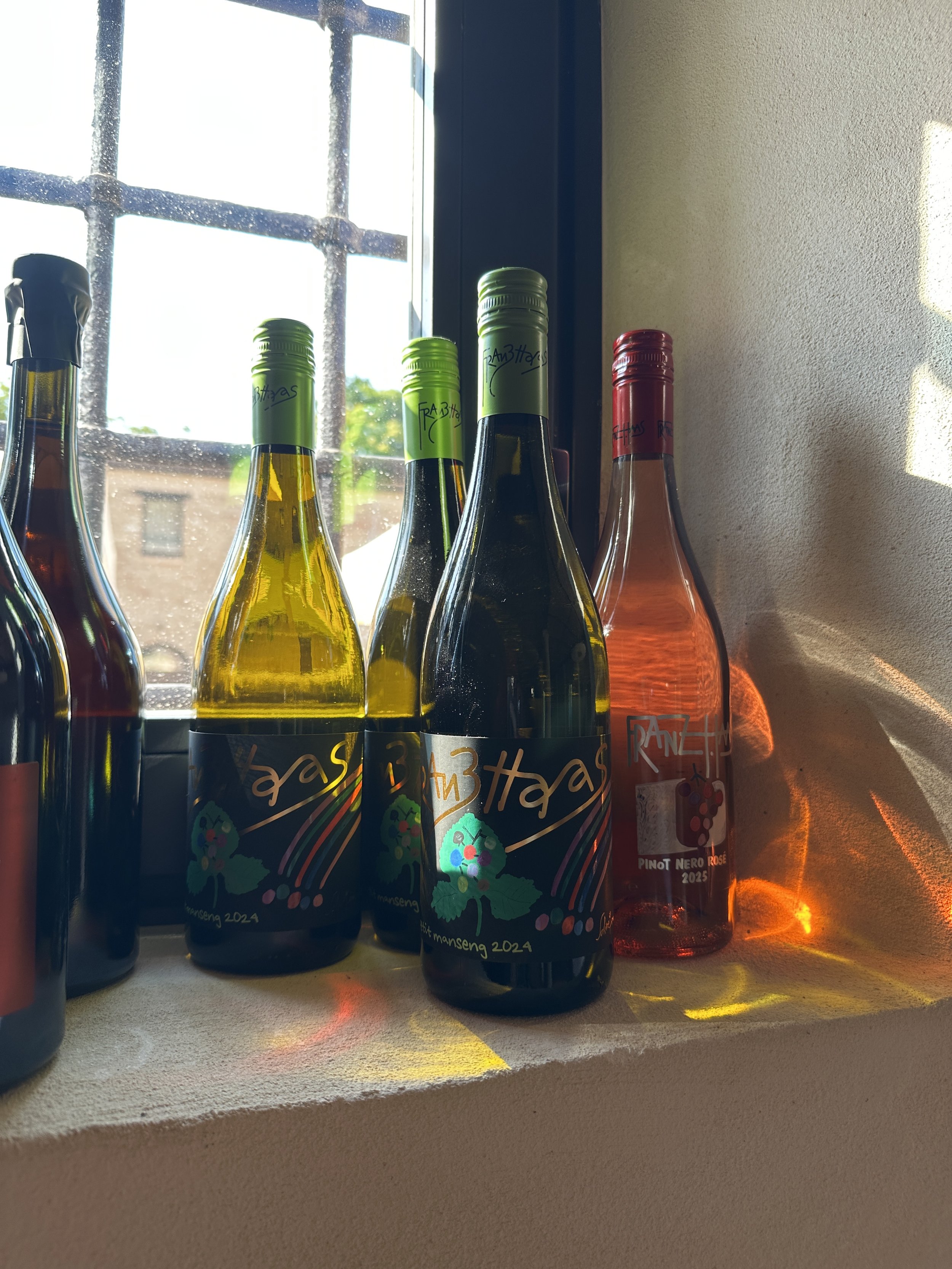 Bottiglie di vino adagiate su una finestra con luce naturale, alcune con etichette colorate e scritte a mano, alcune con etichetta rossa.