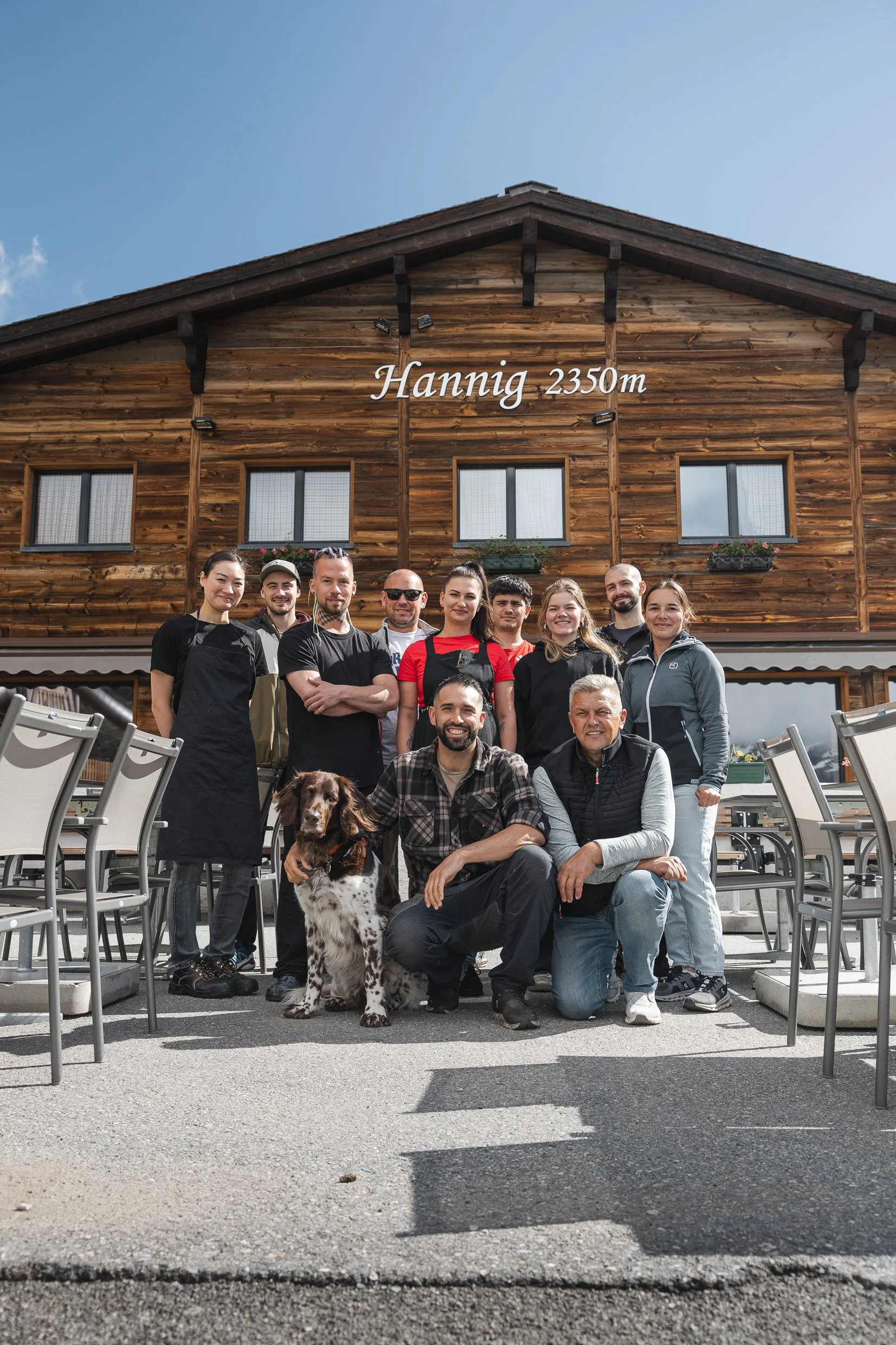 Eine Gruppe von Menschen, die vor einem Holzhaus namens Hannig auf 2350 Metern Höhe stehen, posieren für ein Foto, mit einem Hund in der Nähe.