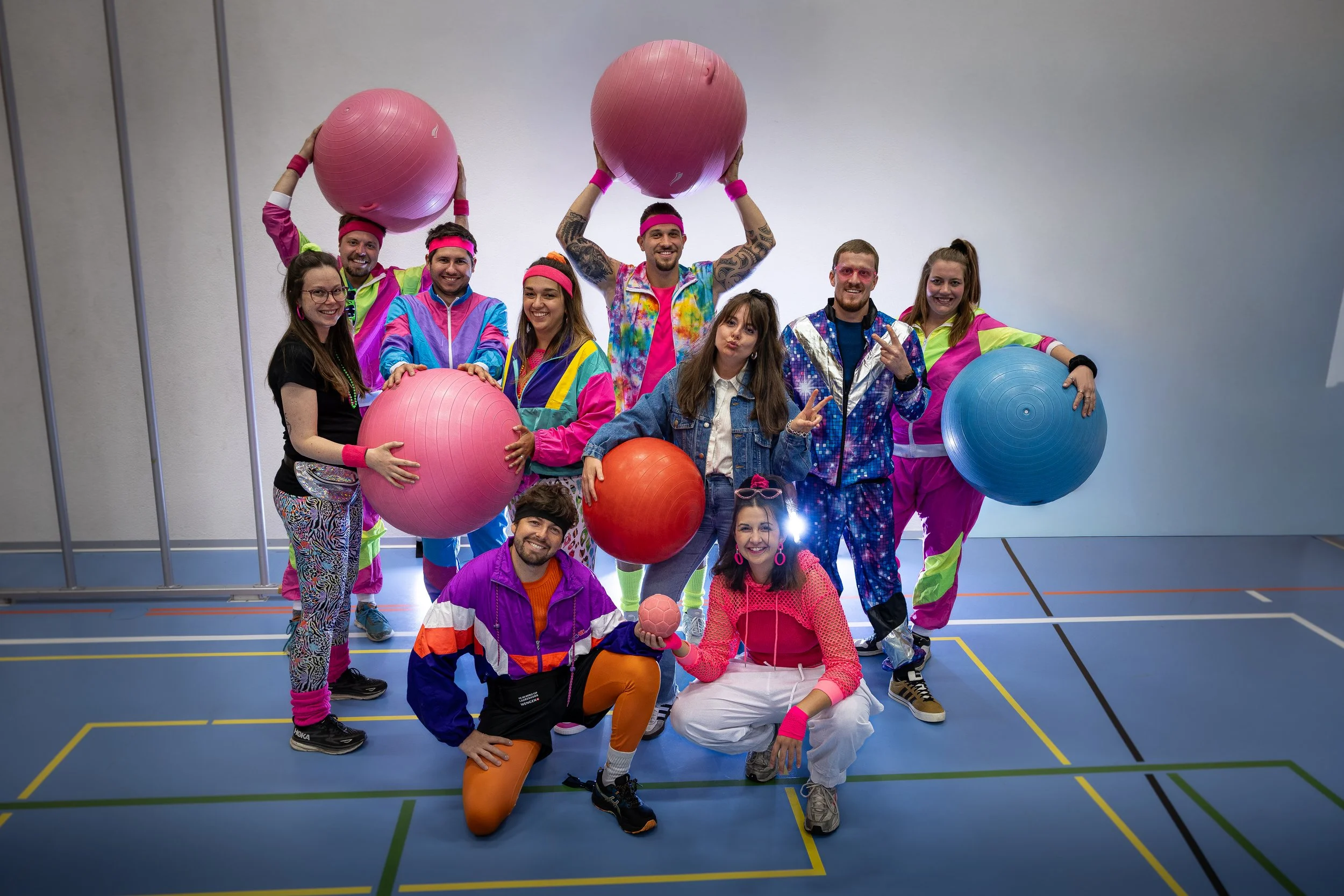 Gruppe von Menschen in bunten 80er-Jahre-Outfits, die sportlich mit großen Gymnastikballen posieren in einer Turnhalle.