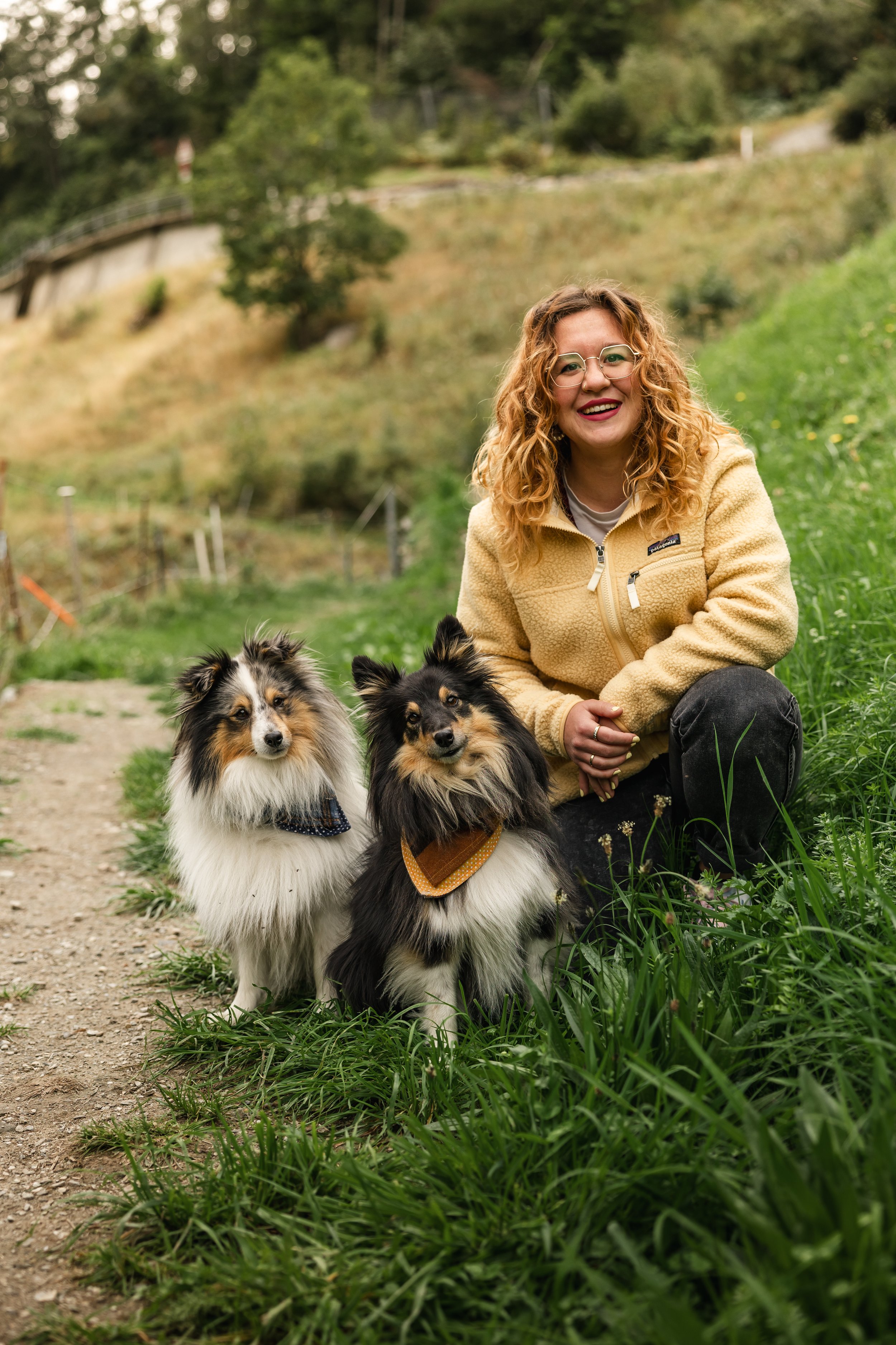 Eine Frau mit lockigem roten Haar, Brille und gelber Jacke sitzt im Grünen, umgeben von zwei Shetland Sheepdogs.