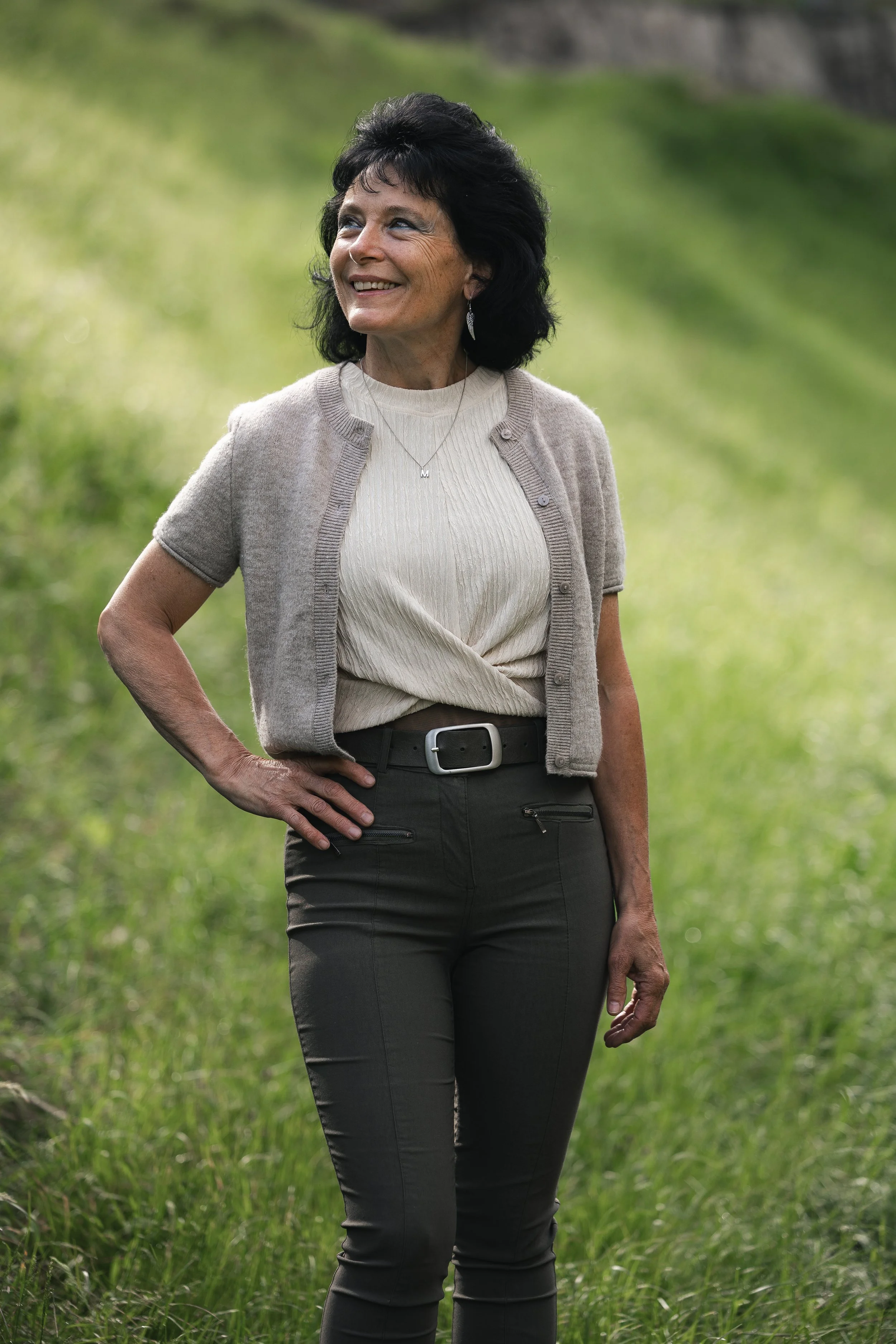 Eine schwarze Frau mit schwarzen Haaren und einem Lächeln im Grünen, trägt eine beige Strickjacke, eine cremefarbene Bluse, schwarze Hose, eine Halskette mit einem Buchstaben "M" und Ohrringe.
