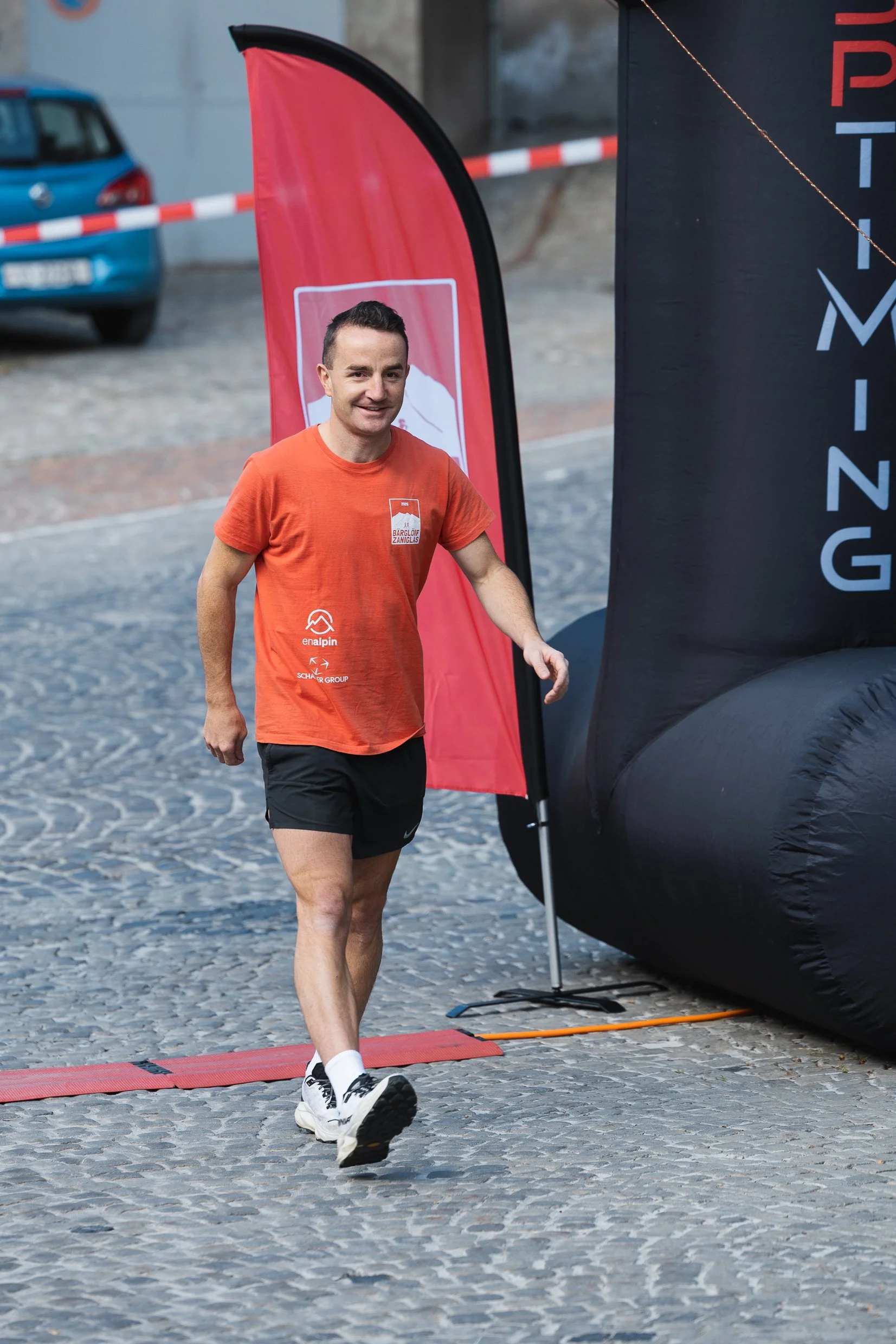 Ein Mann beim Laufen, trägt ein orangefarbenes T-Shirt und schwarze Shorts, am Ziel beim sportlichen Event.