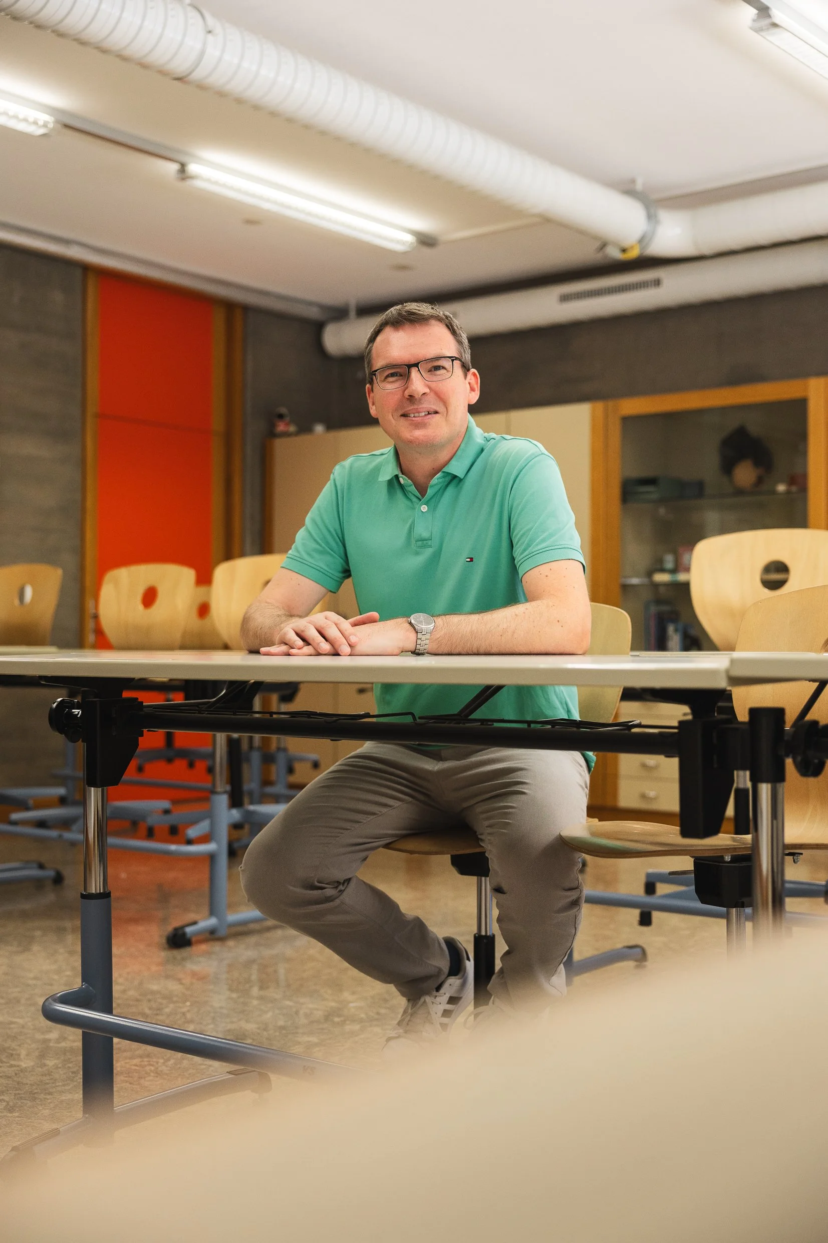 Ein Mann mit Brille trägt ein grünes Polo-Shirt und sitzt an einem Tisch in einem modernen Raum mit Holz und grauen Wänden. Er lächelt in die Kamera.