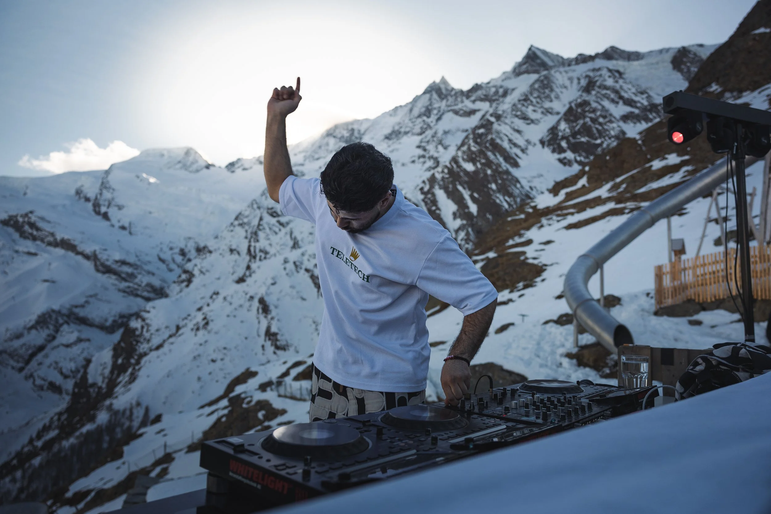 Ein Mann mit dunklem Haar und Brille, der auf einer Berghütte in den Bergen mit Schneeflächen auflegt und auf einem DJ-Controller spielt, während im Hintergrund schneebedeckte Berge zu sehen sind.