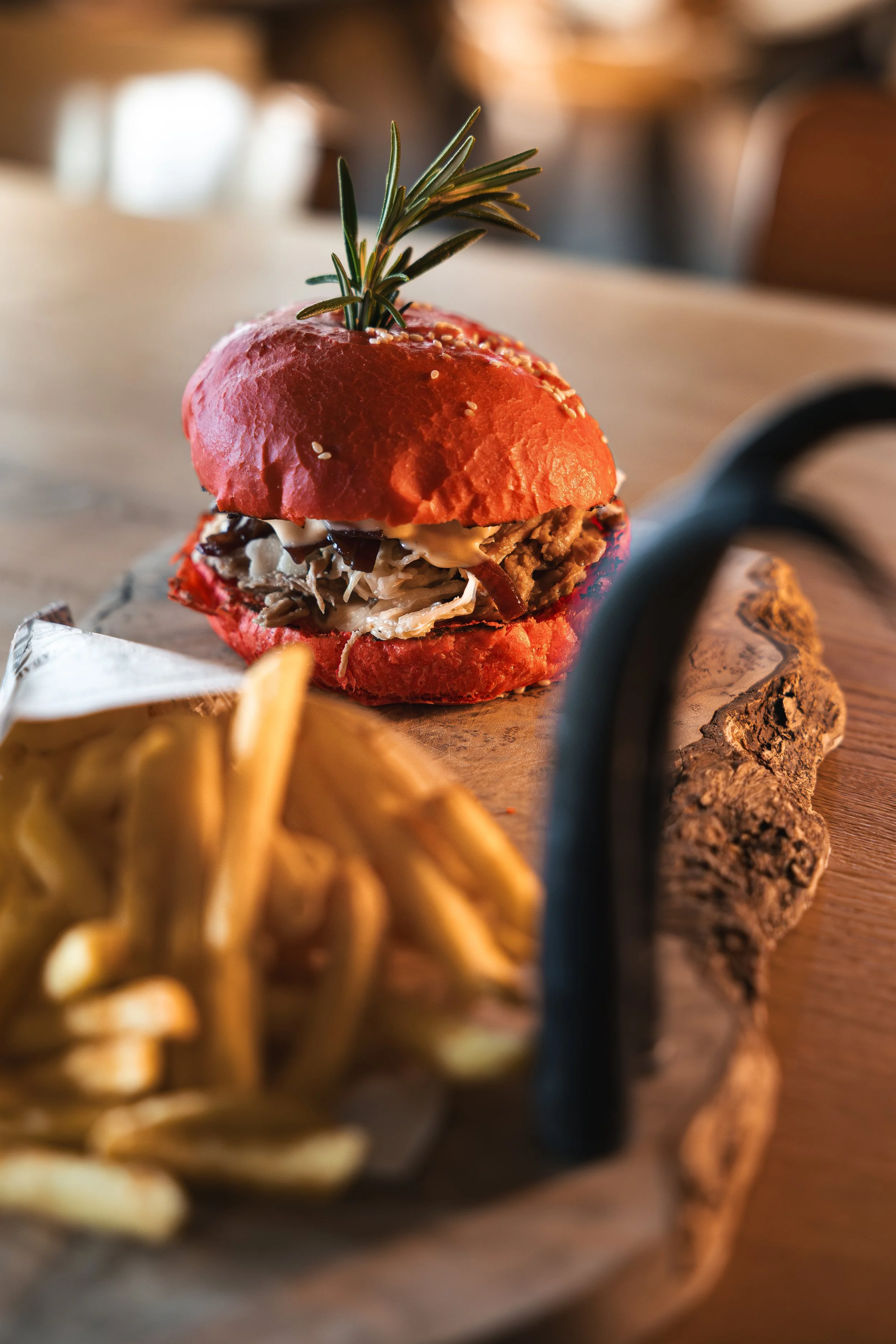Nahaufnahme eines Sandwichs mit rotem Brötchen, gefüllt mit Fleisch und Sauerkraut, garniert mit Rosmarin, auf einem Holzbrett. Im Vordergrund Pommes frites.