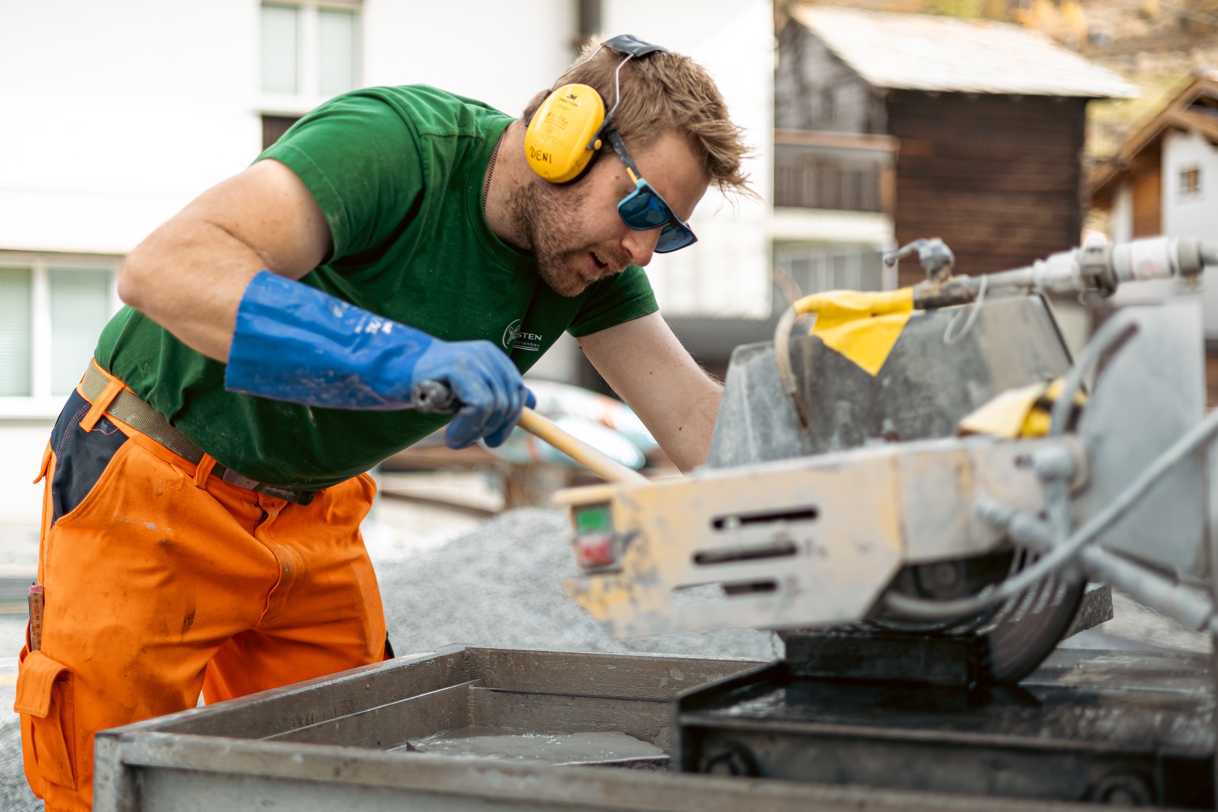Mann beim Beton gießen auf einer Baustelle, trägt orange Arbeitskleidung, grünes T-Shirt und blaue Handschuhe, trägt Gehörschutz und Sonnenbrille.