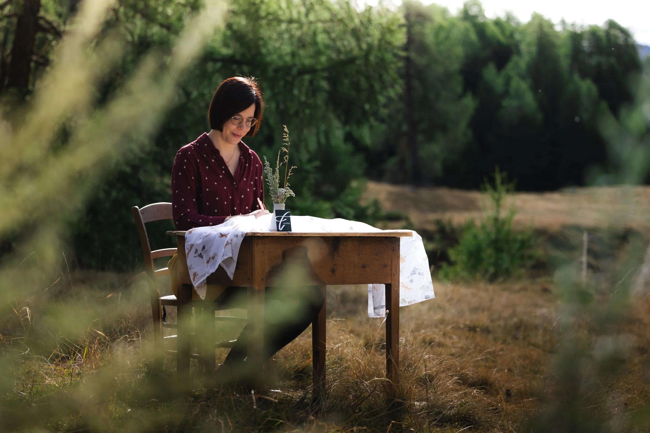 Eine Frau mit dunklem Haarschnitt und Brille sitzt an einem Holztisch im Freien, umgeben von Bäumen, bei Sonnenlicht, mit einer weißen Tischdecke und einer Vase mit Pflanzen auf dem Tisch.