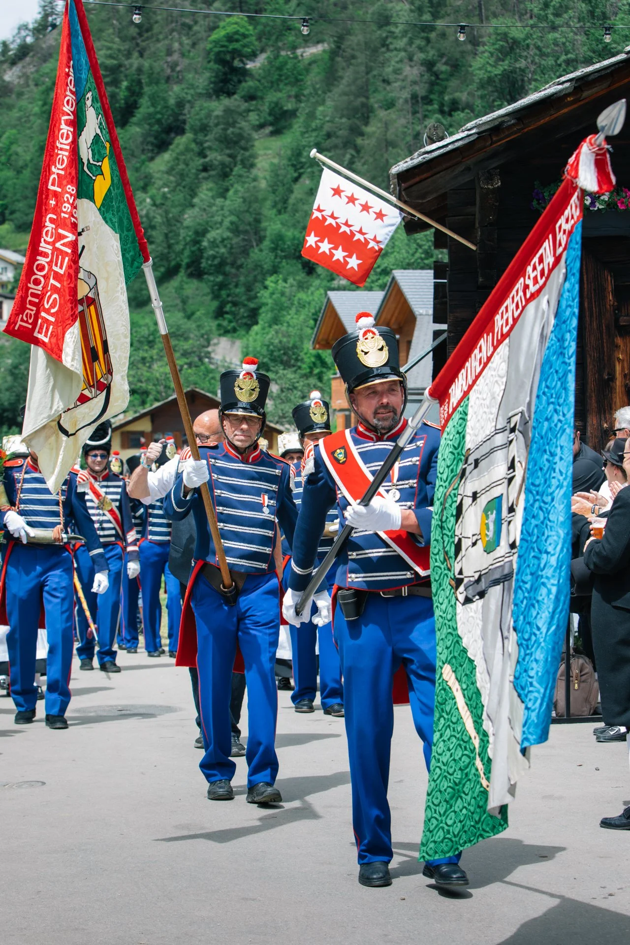 Musiker in historischen Uniformen bei einem Festumzug, mit Fahnen und Präsentationsbannern, in einer malerischen Gegend mit Hügeln und Holzhäusern.