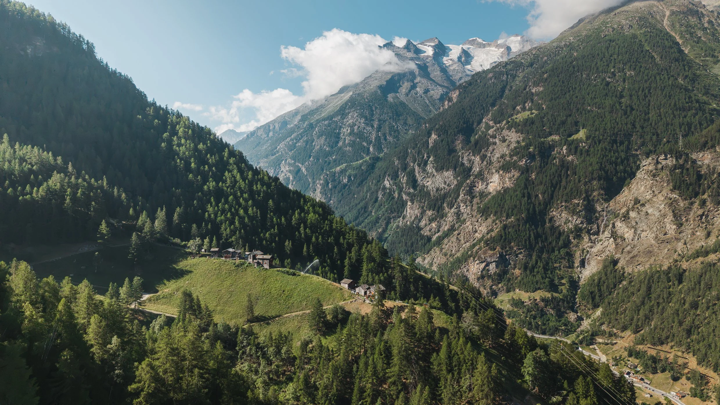 Eine Berglandschaft mit grünen Wäldern, zerstreuten Häusern im Tal und hohen schneebedeckten Gipfeln im Hintergrund, umgeben von Wolken und blauem Himmel.