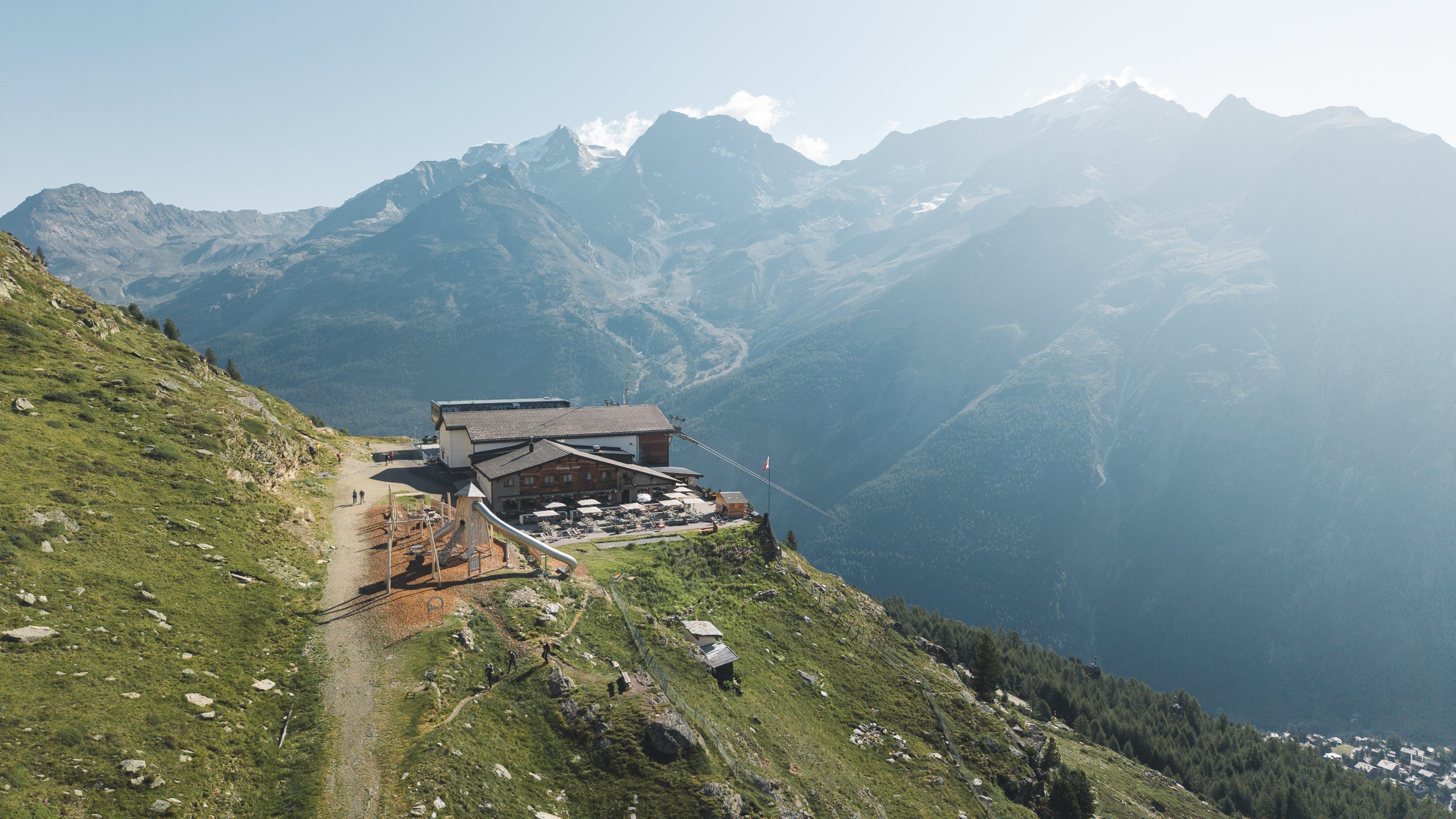 Hütte auf einer bergigen Landschaft mit Blick auf hohe, schneebedeckte Berge und eine Seilbahn in der Ferne.