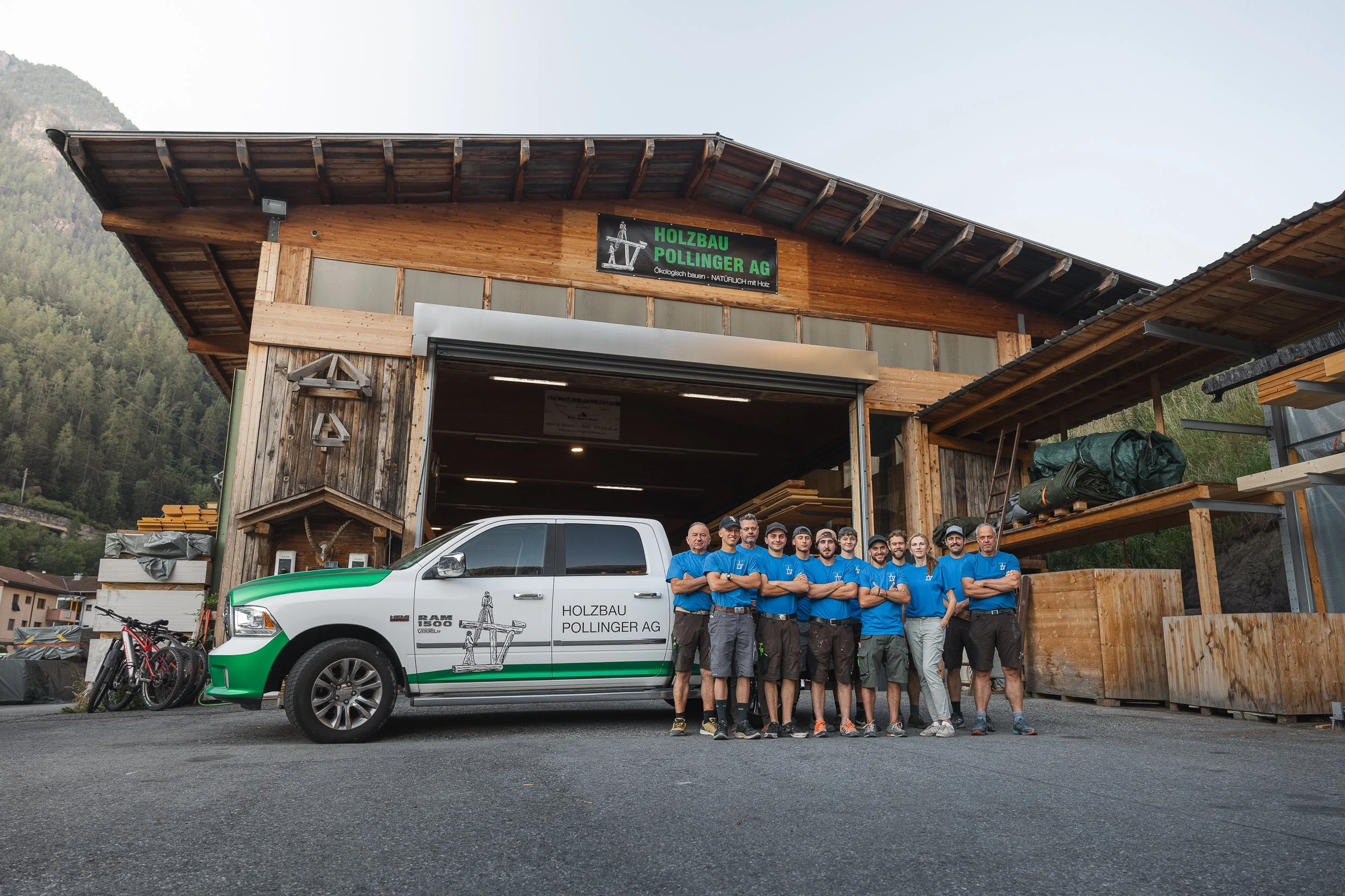 Gruppenfoto von Mitarbeitern der Holzbau Pollinger AG vor einem Holzbau-Unternehmen, mit einem weißen Pickup mit Firmenlogo, in einer montierten Holzstruktur im Hintergrund, umgeben von Bergen, bei sonnigem Wetter.