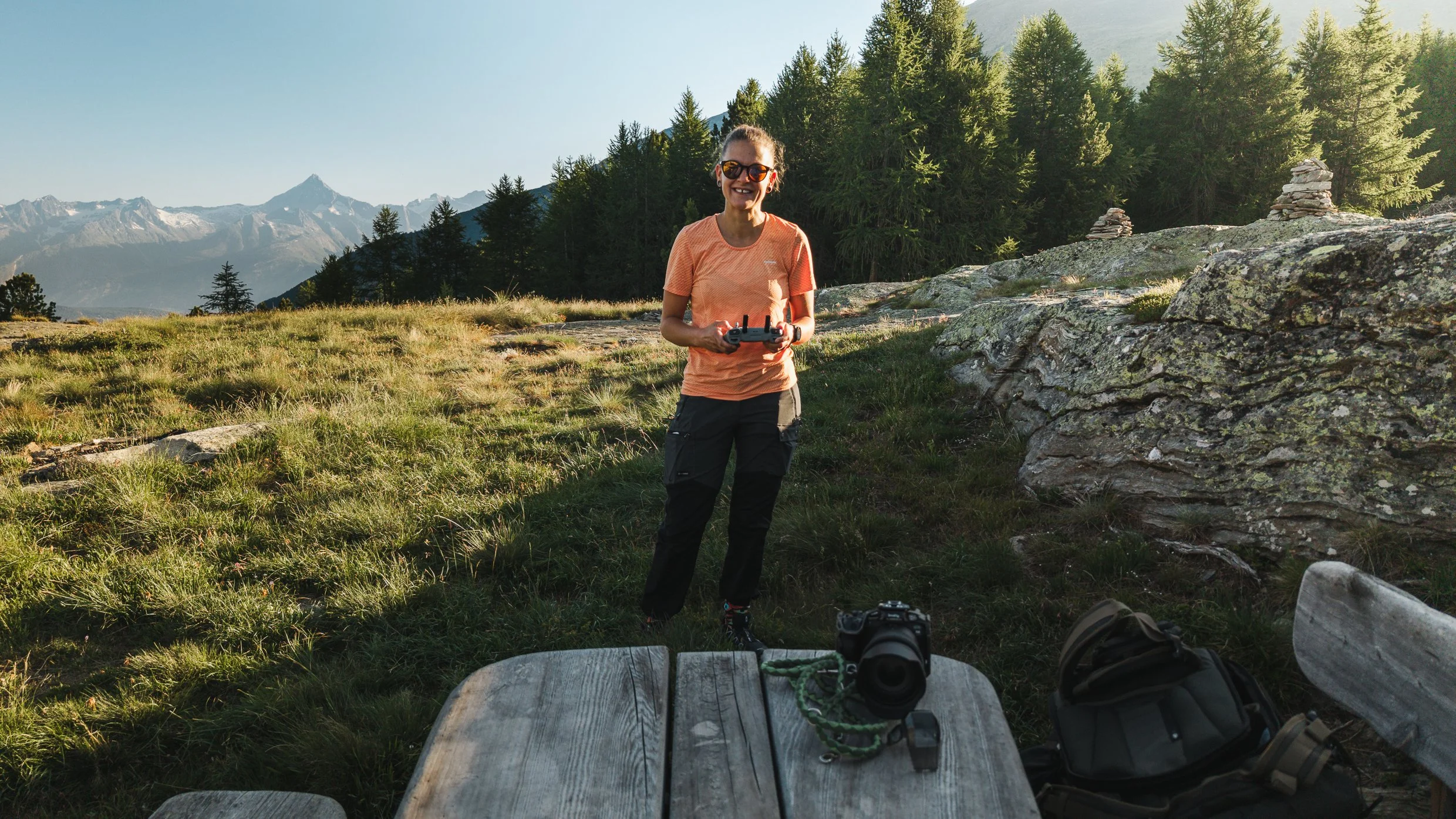 Eine lachende Frau mit Sonnenbrille hält eine Fernbedienung in der Hand, in einer Berglandschaft mit Tannenbäumen und Bergen im Hintergrund, beim Drohnenflug.