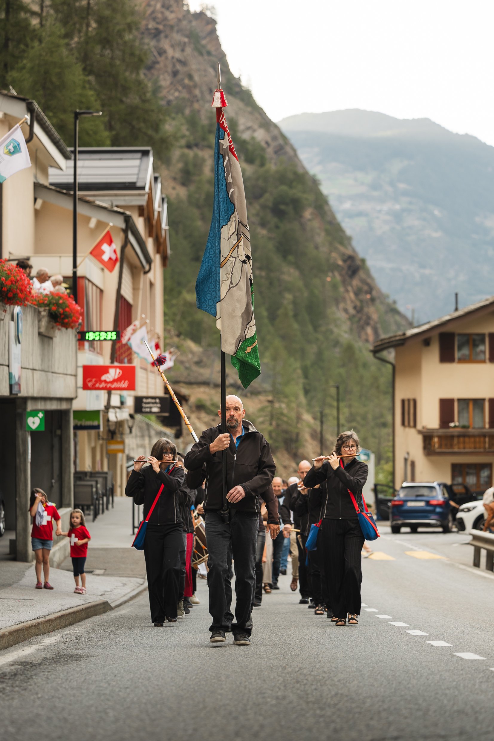 Menschenpartizipation bei einer Parade in einer kleinen Stadt mit Bergen im Hintergrund, einige Teilnehmer tragen traditionell Schweizer Kleidung und halten Flaggen. Es sind sowohl Erwachsene als auch Kinder beteiligt.