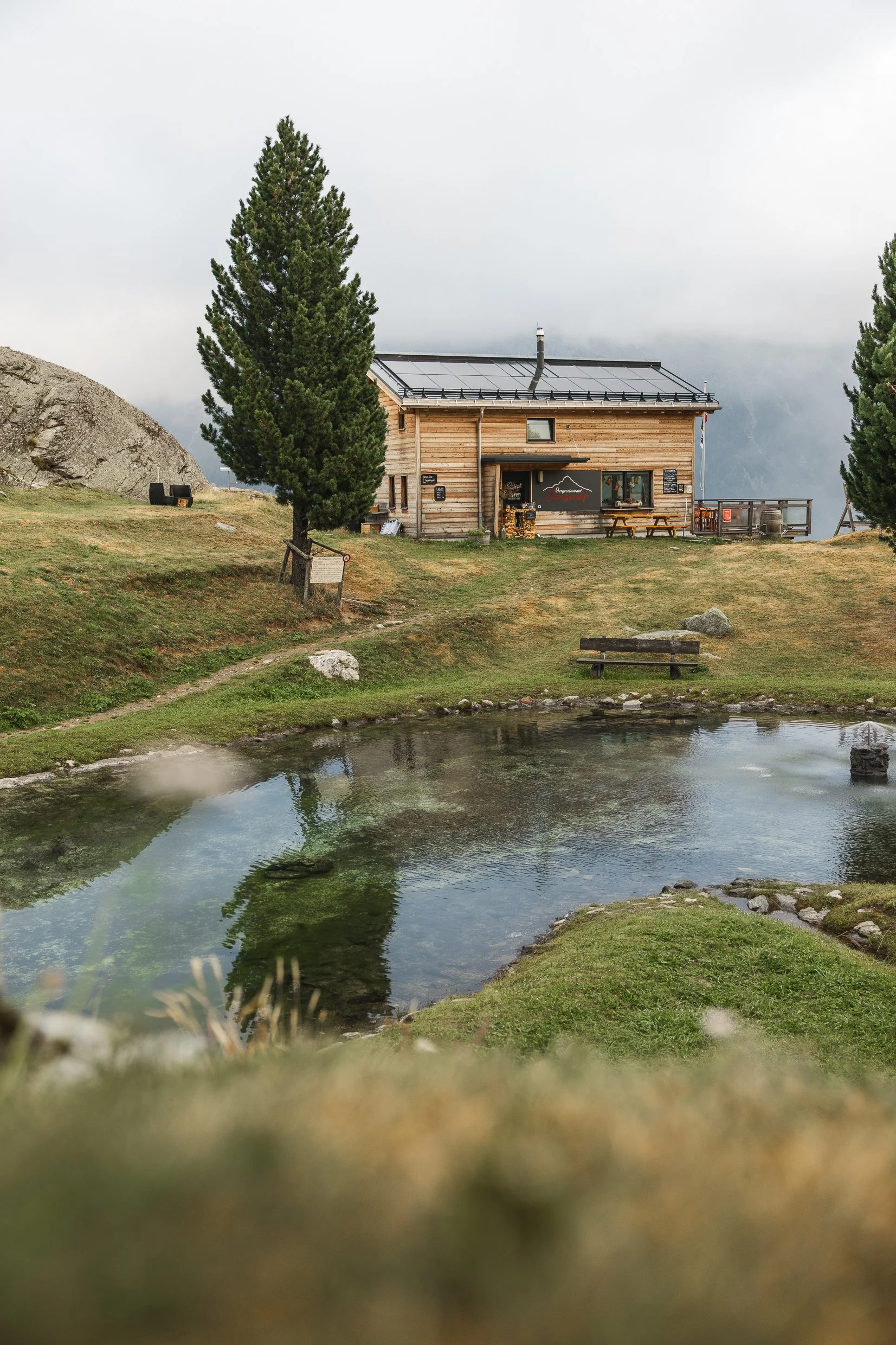 Ein holzgastr der Berghütte mit Tischen, Bänken und einer Sonnenterrasse, umgeben von Bäumen, inmitten einer grünen Landschaft mit einem kleinen Wasserfall im Vordergrund, Berge und wolkiger Himmel im Hintergrund.