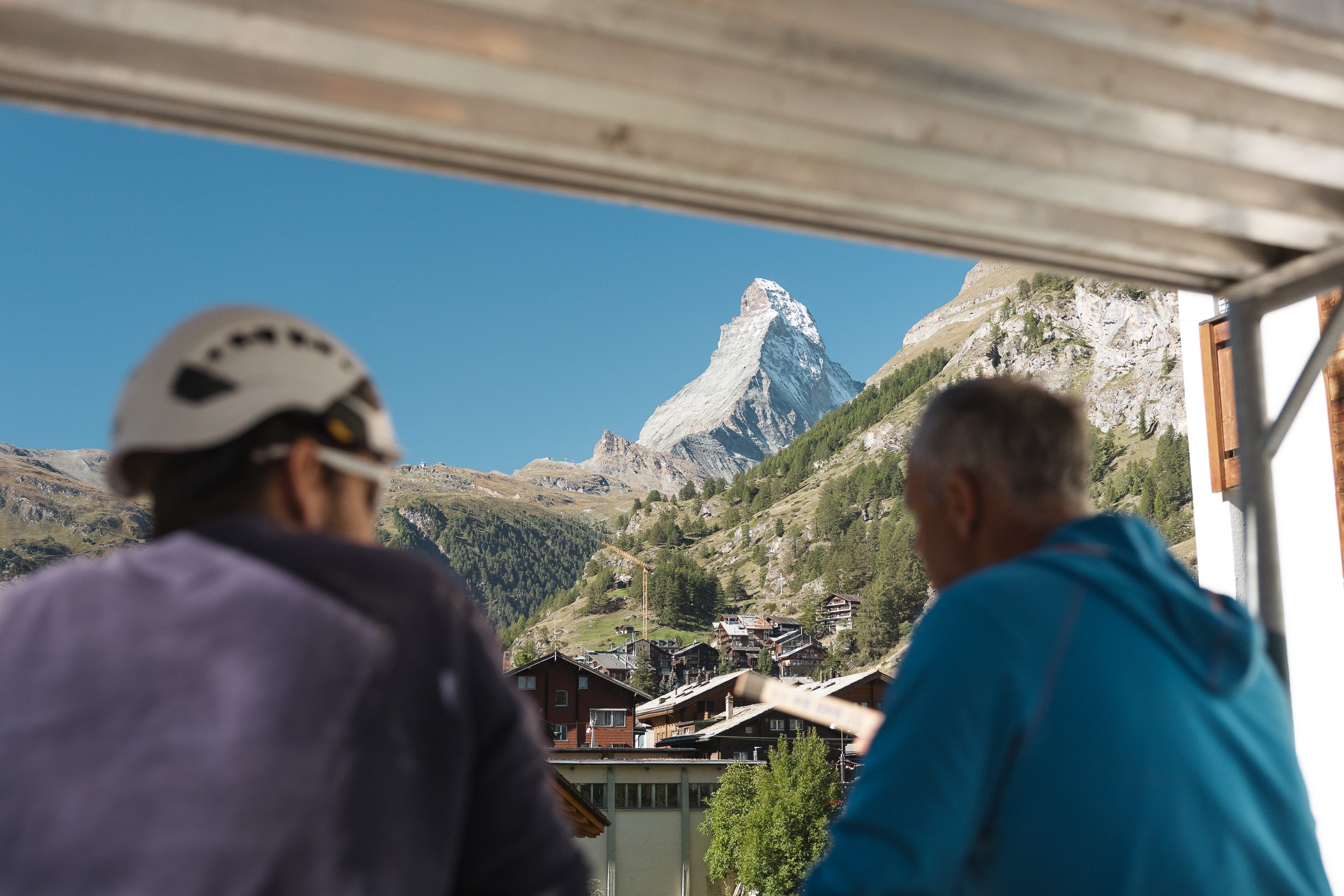Blick auf die Schneekuppe des Matterhorns, umrahmt von grünen Hügeln und einer kleinen Ortschaft, teilweise blickt man durch eine offene Fenster- oder Türöffnung.