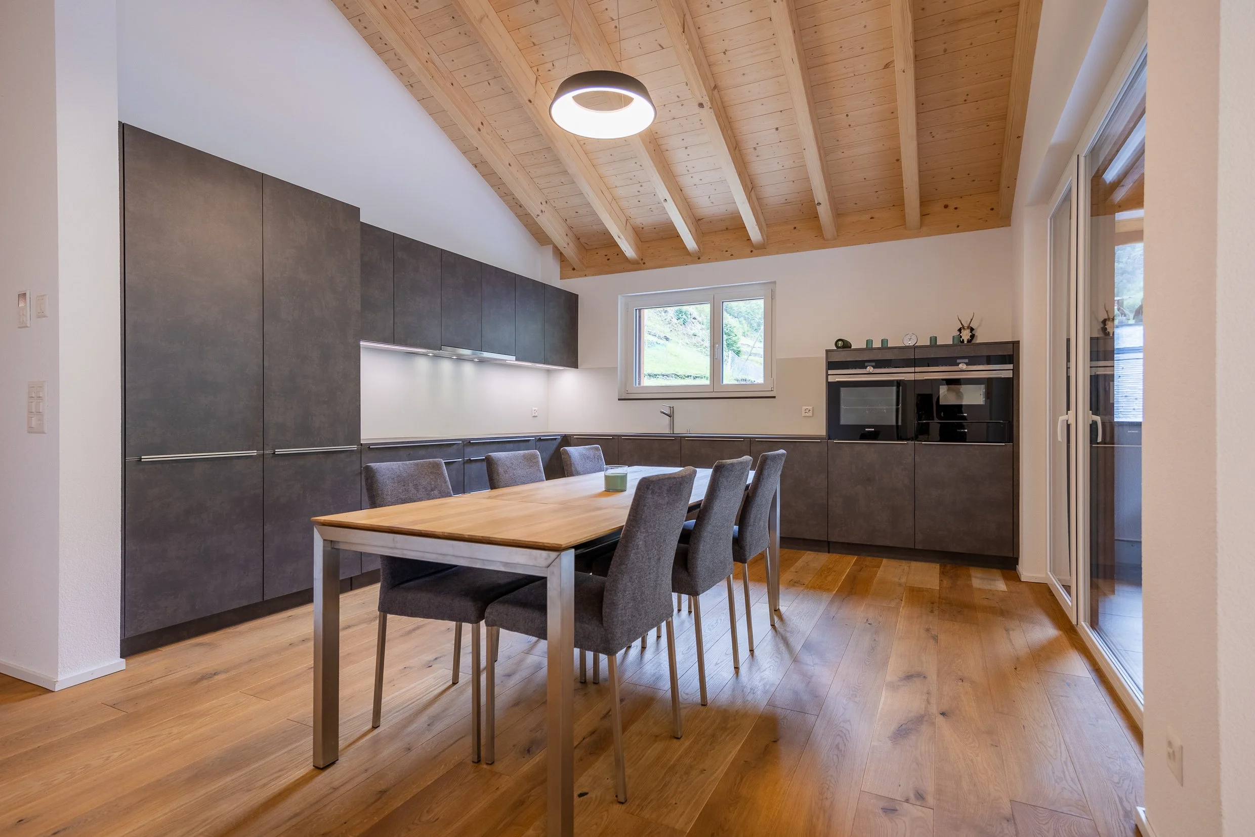 Moderne Küche mit Holzdecke, grauen Schränken, einen Esstisch und Stühlen, Fenster mit Blick nach draußen, und Deckenleuchte.