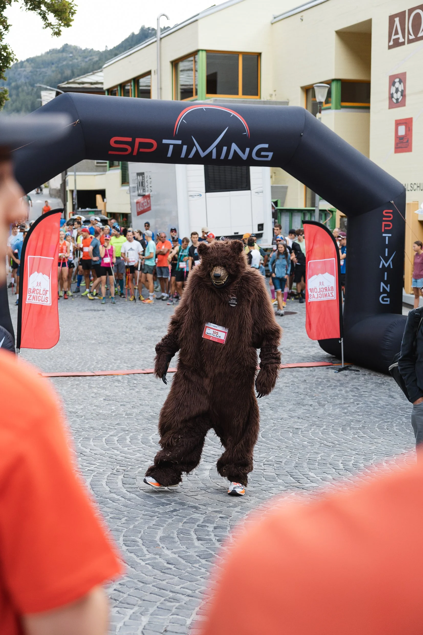 Ein Bärenkostüm bei einem Lauf-Event, im Hintergrund viele Läufer und ein Gebäude, vor dem Bär. Das Bild ist an einem Stadtplatz aufgenommen.