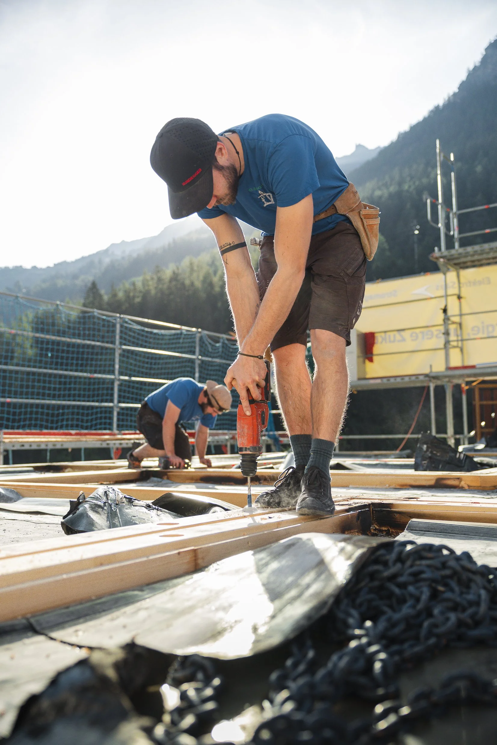 Mann arbeitet mit einem Elektrowerkzeug auf einer Baustelle im Freien, im Hintergrund Berge und eine Wolkendecke, trägt schwarze Schuhe, dunkelbraune Shorts, blauen T-Shirt, schwarze Cap, und hat Tattoos am Unterarm.