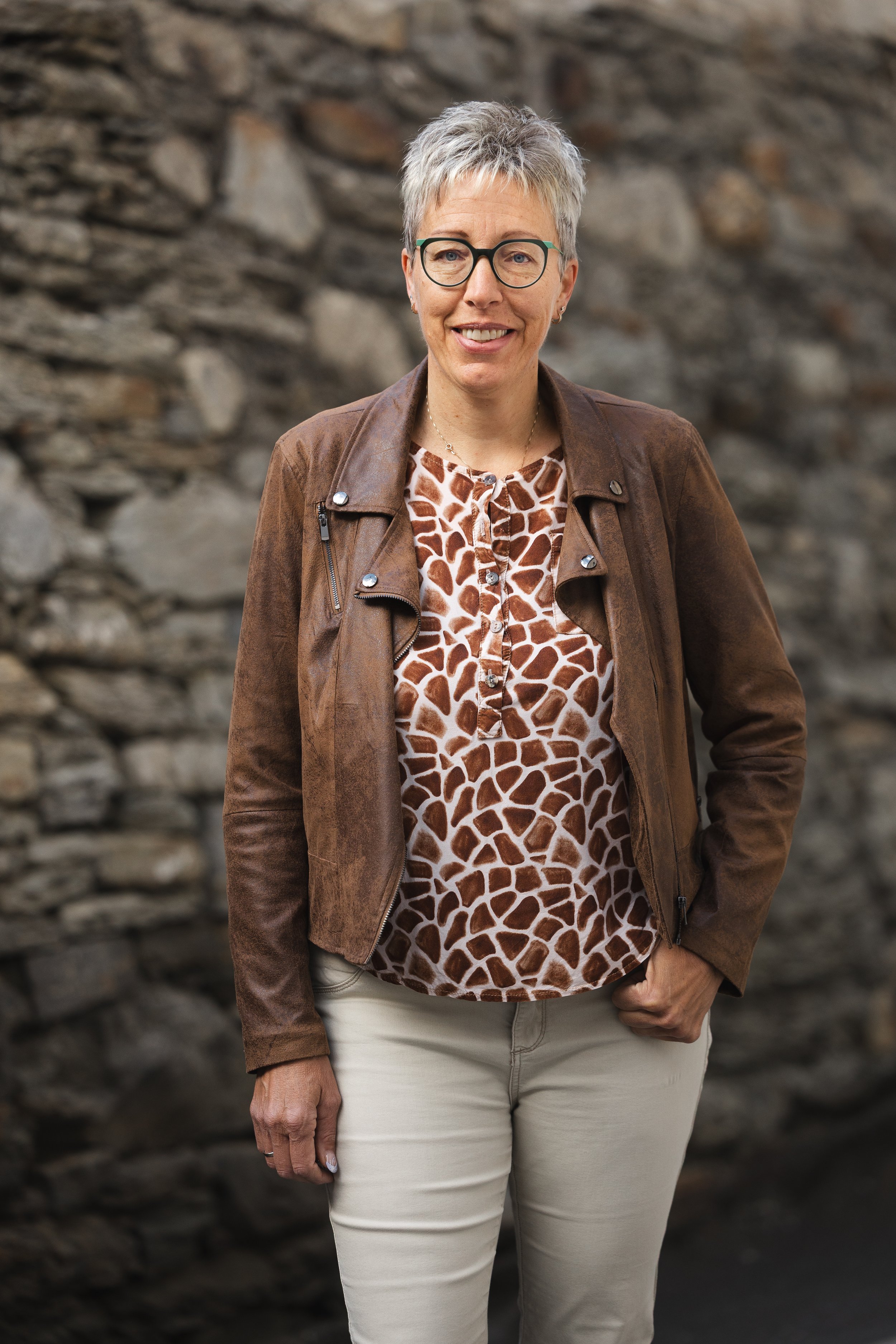 Eine älter Frau, die Brille trägt, trägt eine braune Lederjacke, ein Hemd mit Giraffenmuster, und beige Hose. Sie steht vor einer Steinmauer und lächelt.