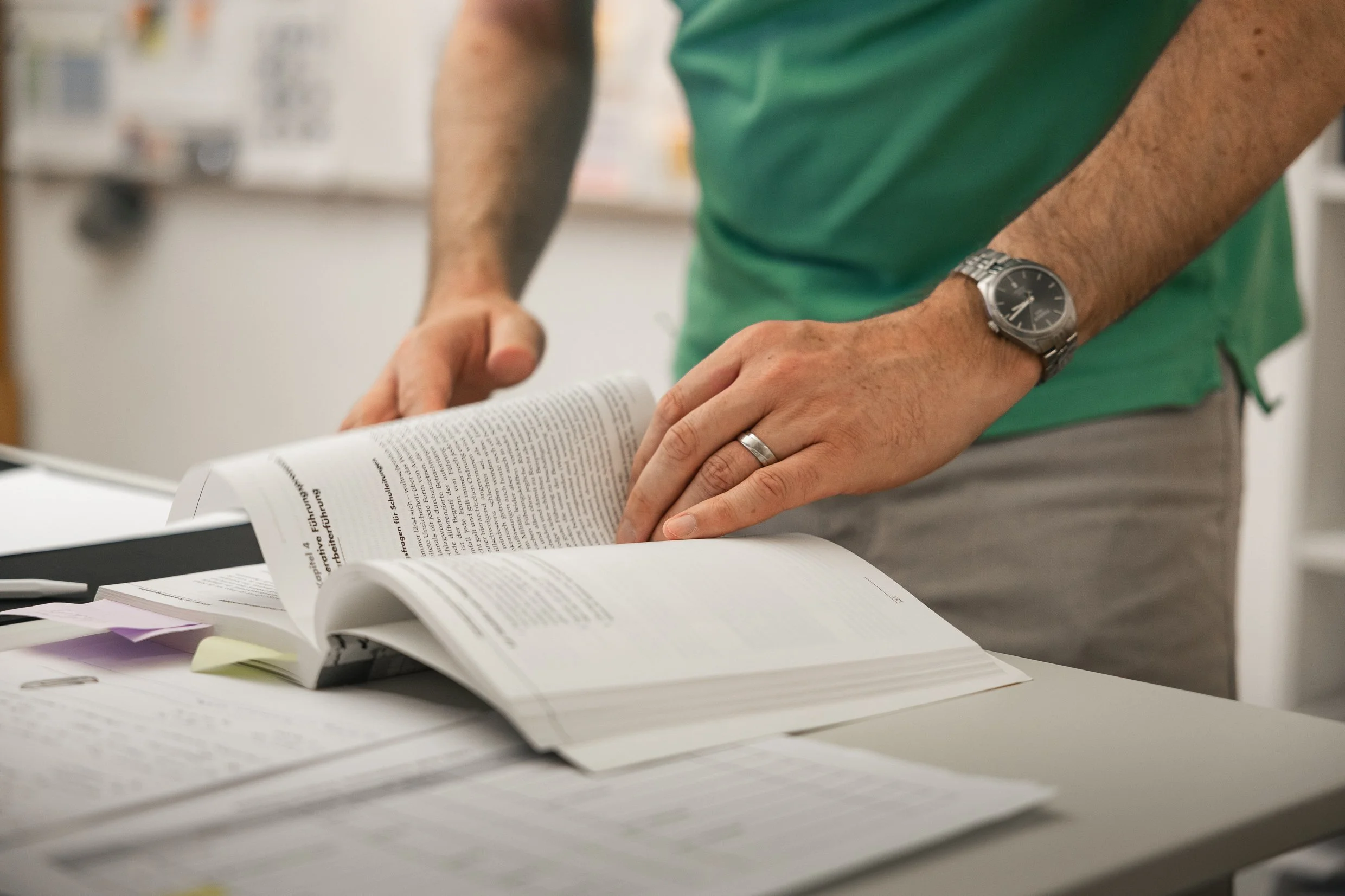 Person in grünem T-Shirt liest in einem geöffneten Buch, auf einem Schreibtisch mit Notizen und Unterlagen. Hände mit Arm und Uhr sichtbar.