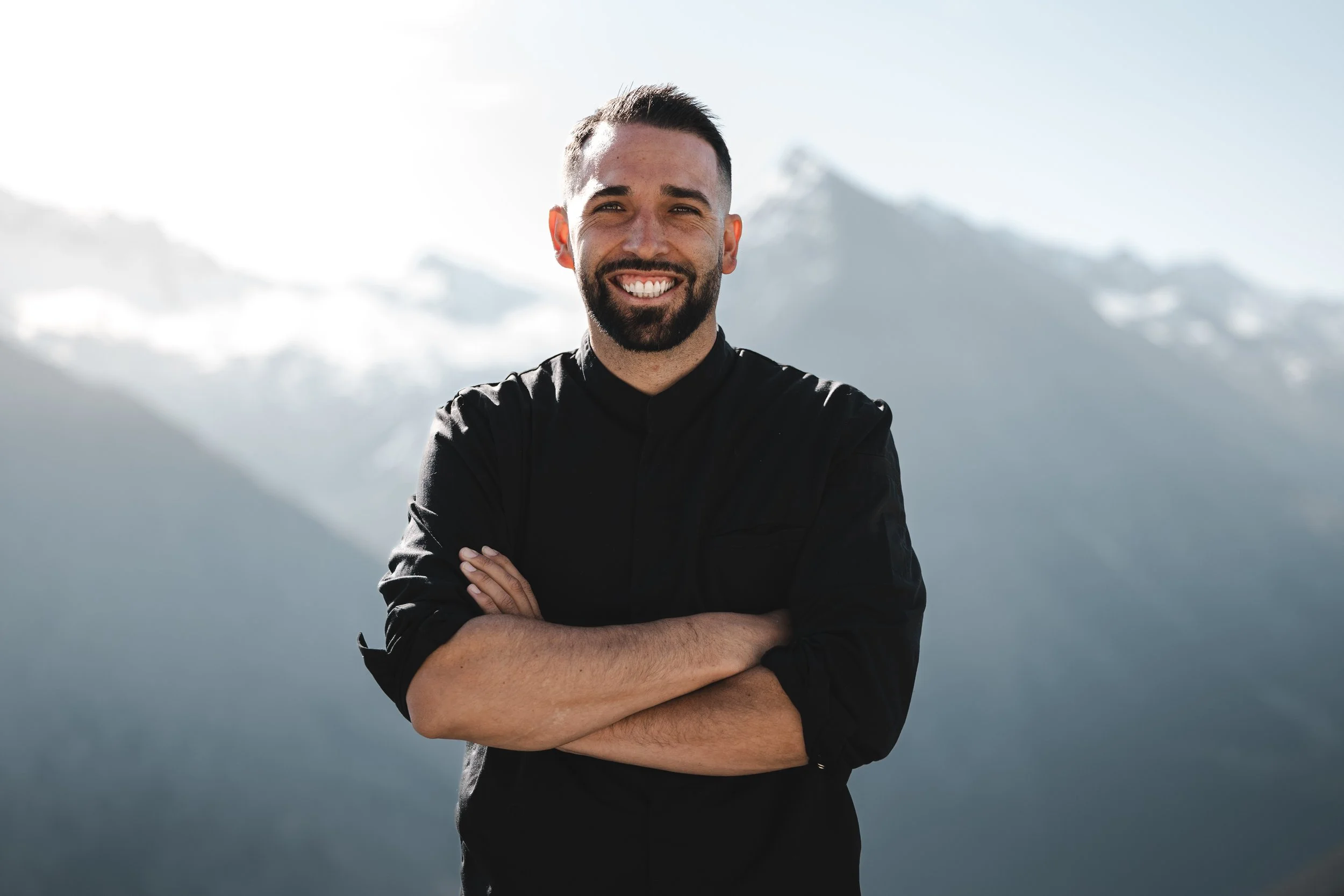 Eine Person in Winterkleidung steht neben einem Feuer im Schnee in einer Berglandschaft bei Dämmerung, mit schneebedeckten Bergen im Hintergrund.