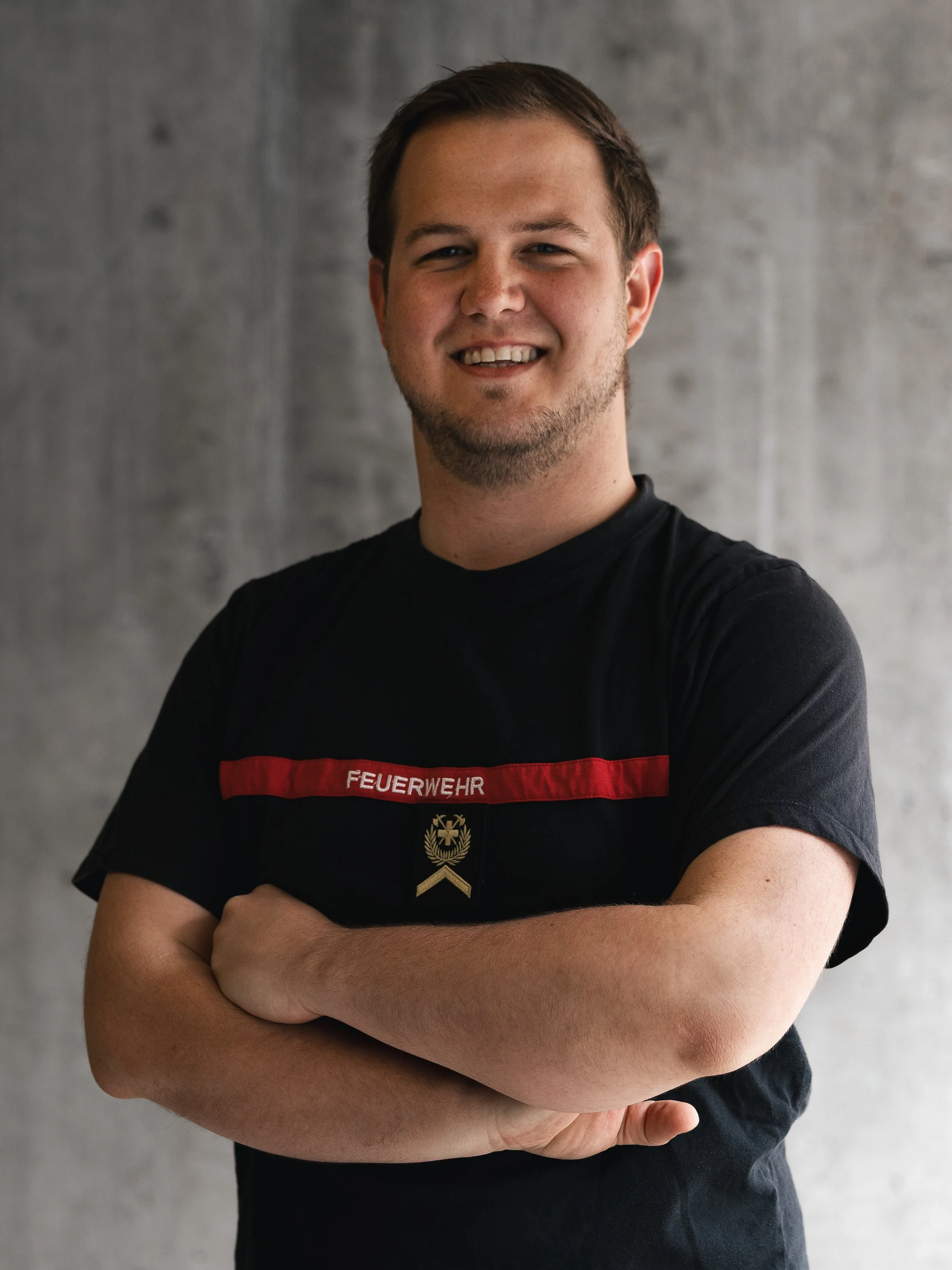 Junger Mann in Feuerwehruniform mit verschränkten Armen, lächelnd vor einer grauen Wand.