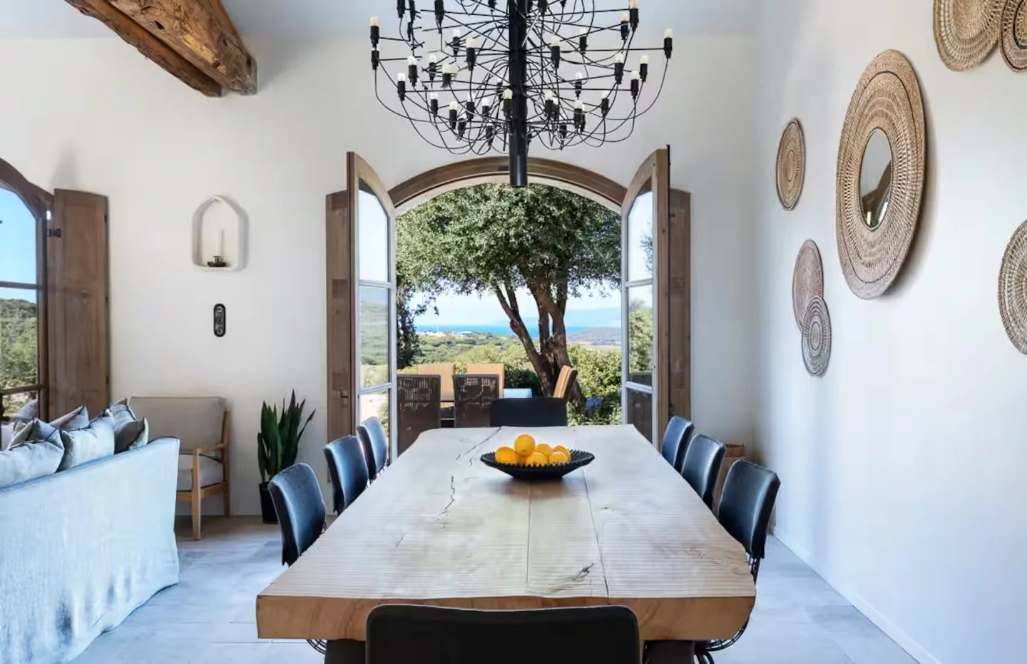 Salle à manger lumineuse avec une grande table en bois et des chaises noires, ouverte sur un jardin avec vue sur des arbres et la mer. Décoration moderne avec miroirs en rotin sur le mur et un chandelier noir.