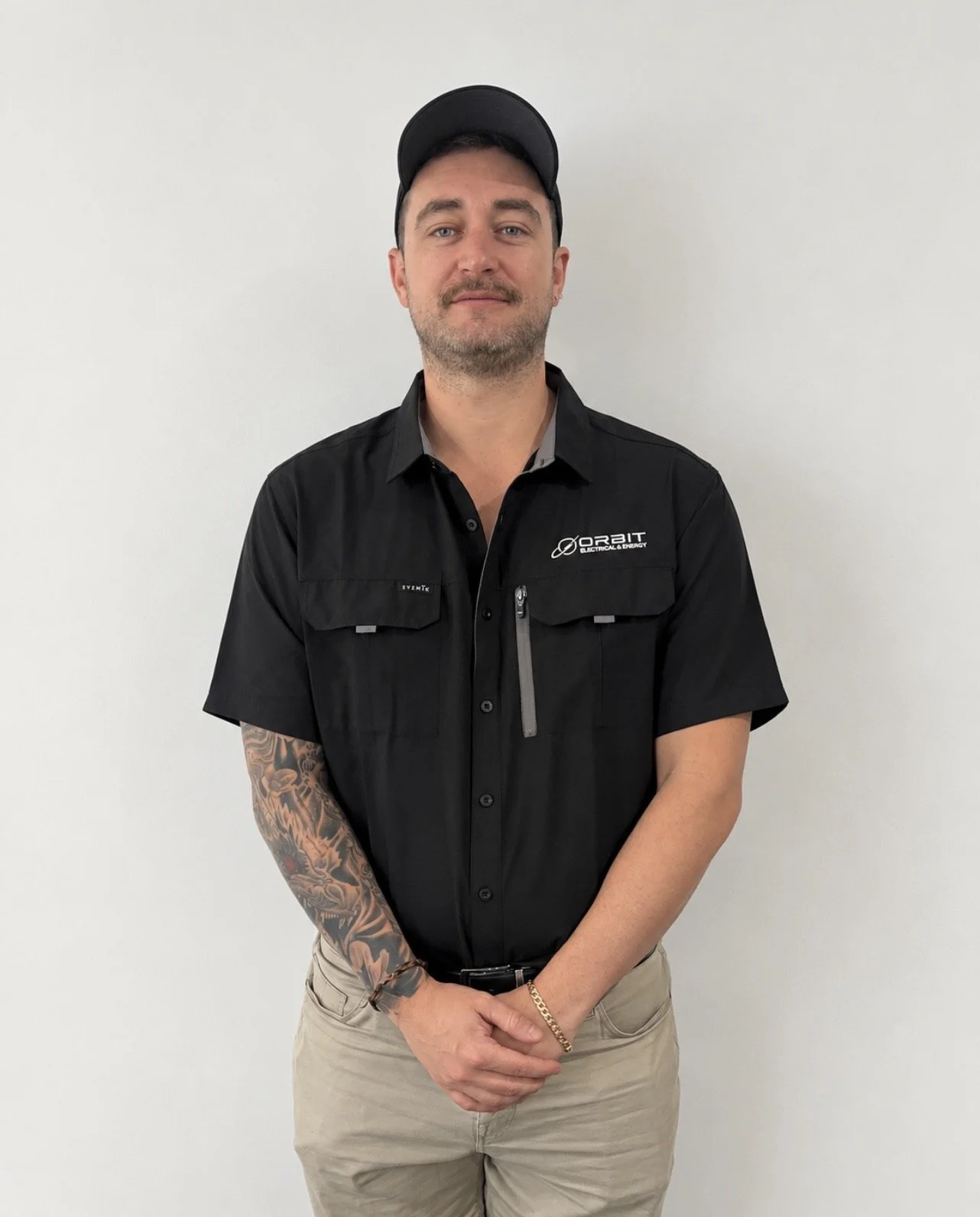 A man with a cap, short beard, wearing a black shirt with company logo, beige pants, and a tattooed arm, standing against a plain light-colored background.