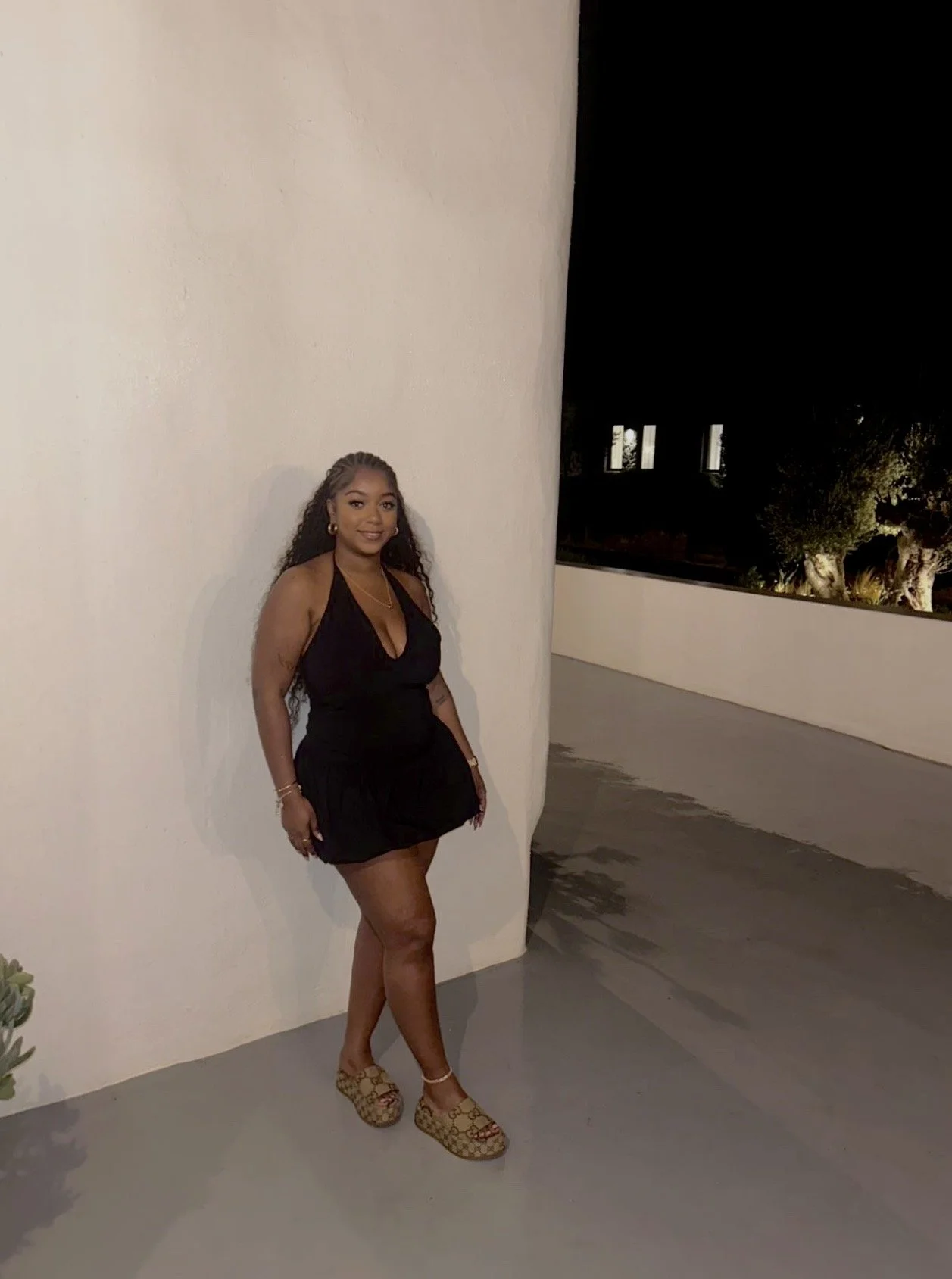 A woman with long curly hair and jewelry wearing a black dress and patterned platform shoes, standing against a white curved wall at night with dark trees and windows in the background.