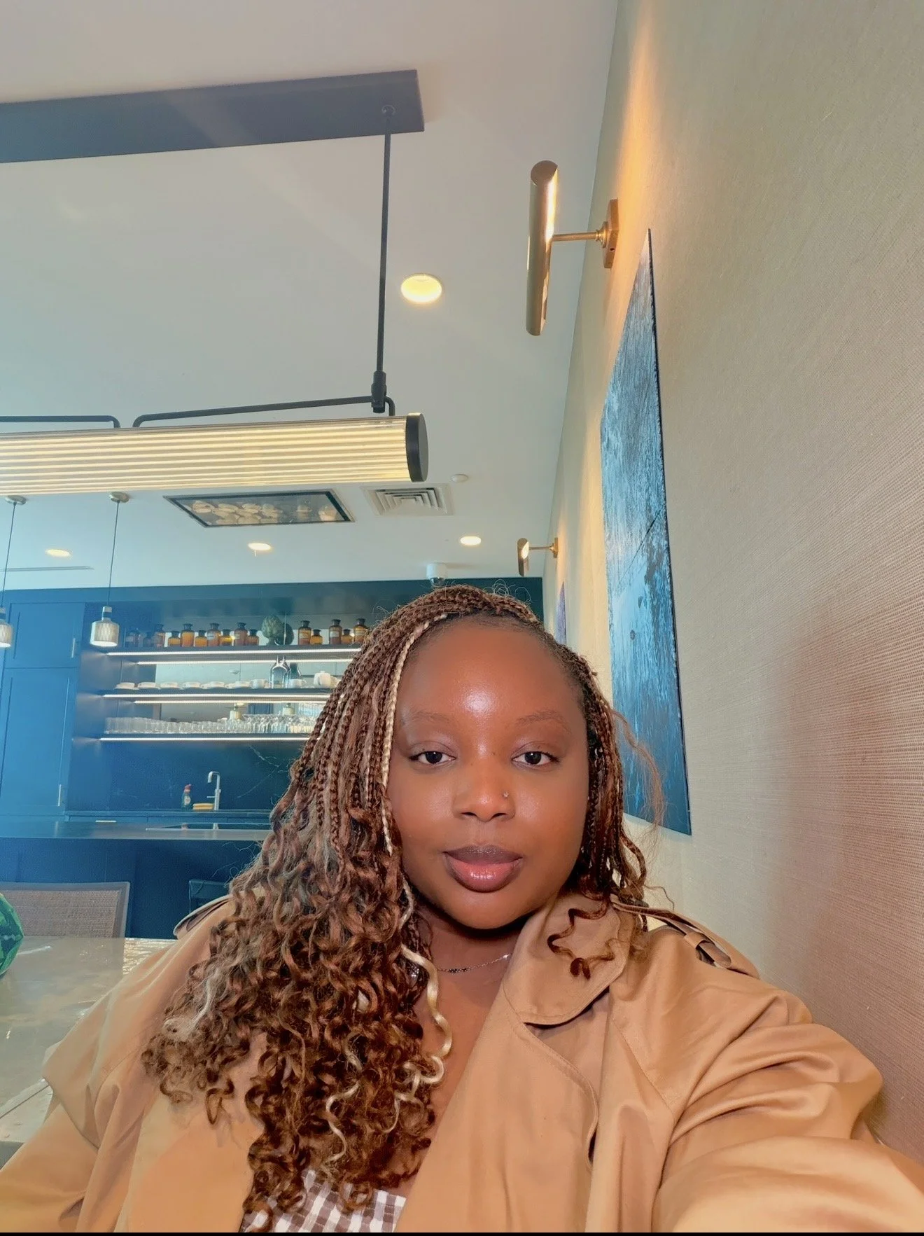 A woman with curly hair sits at a table in a modern restaurant or cafe, with a blue and beige interior and hanging lights in the background.