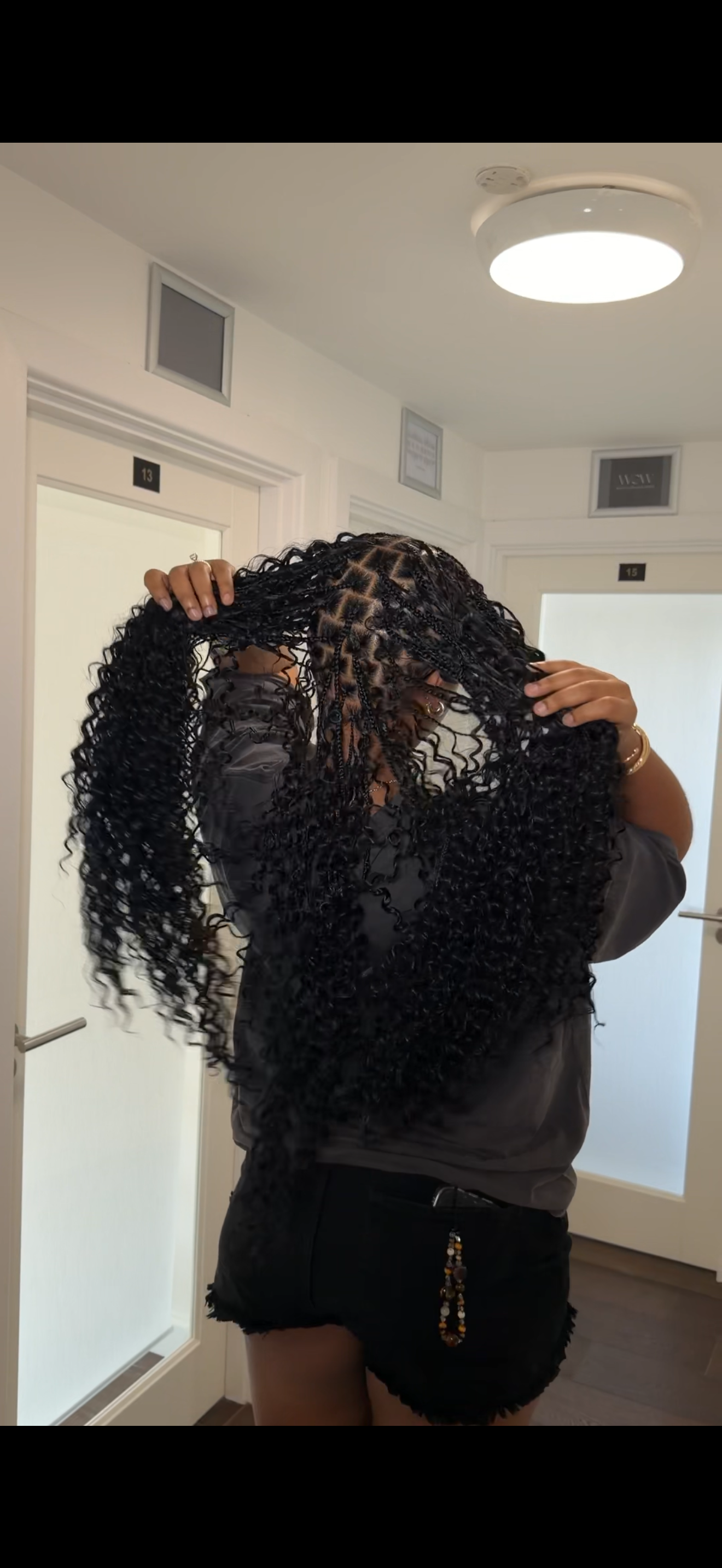 Woman with long, curly braids standing in front of a mirror, holding her hair up in a room with white walls and doors labeled 13 and 15.