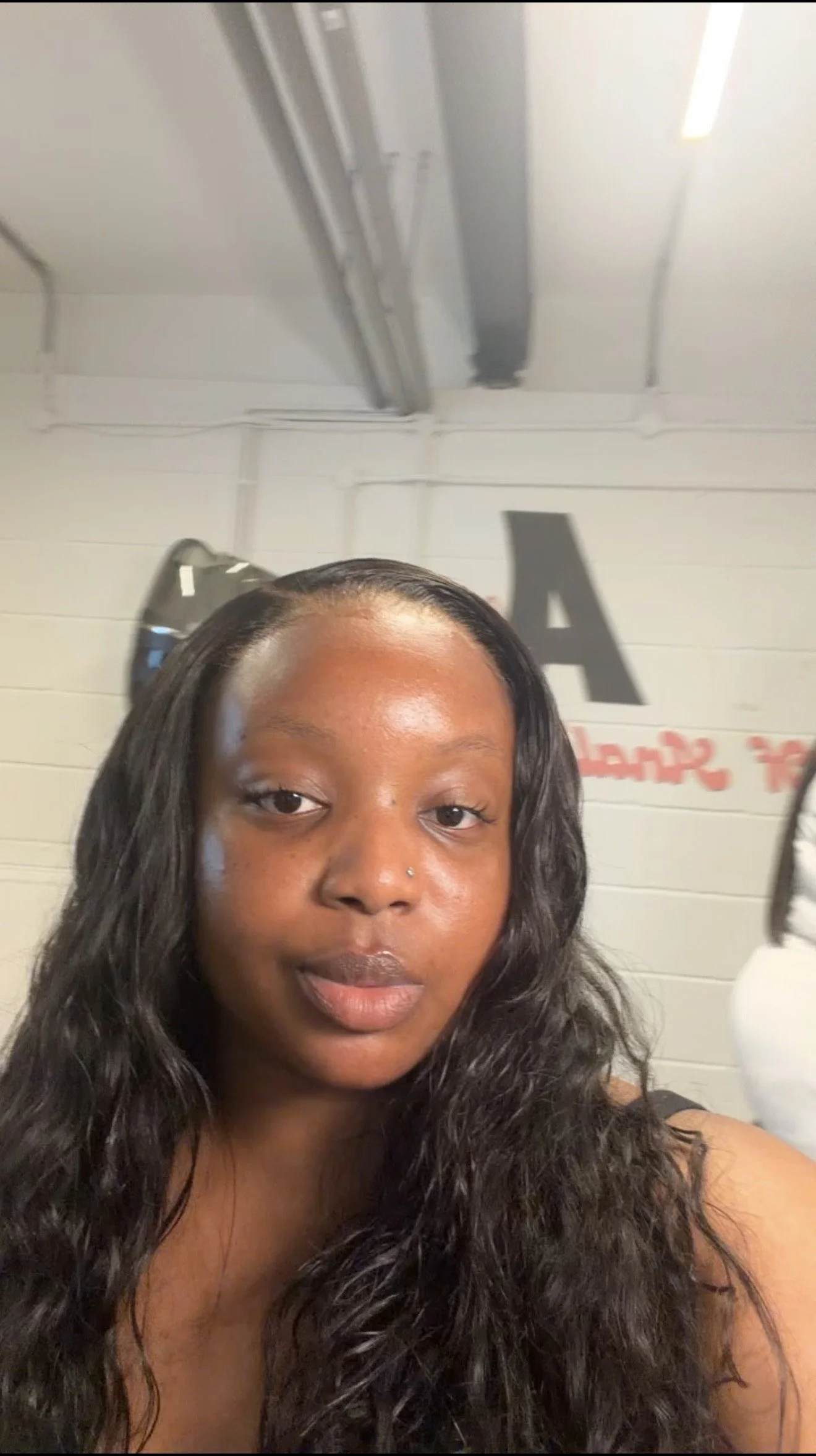 Close-up of a woman with long curly dark hair, wearing a small nose ring, looking at the camera inside a room with white walls and some text in the background.