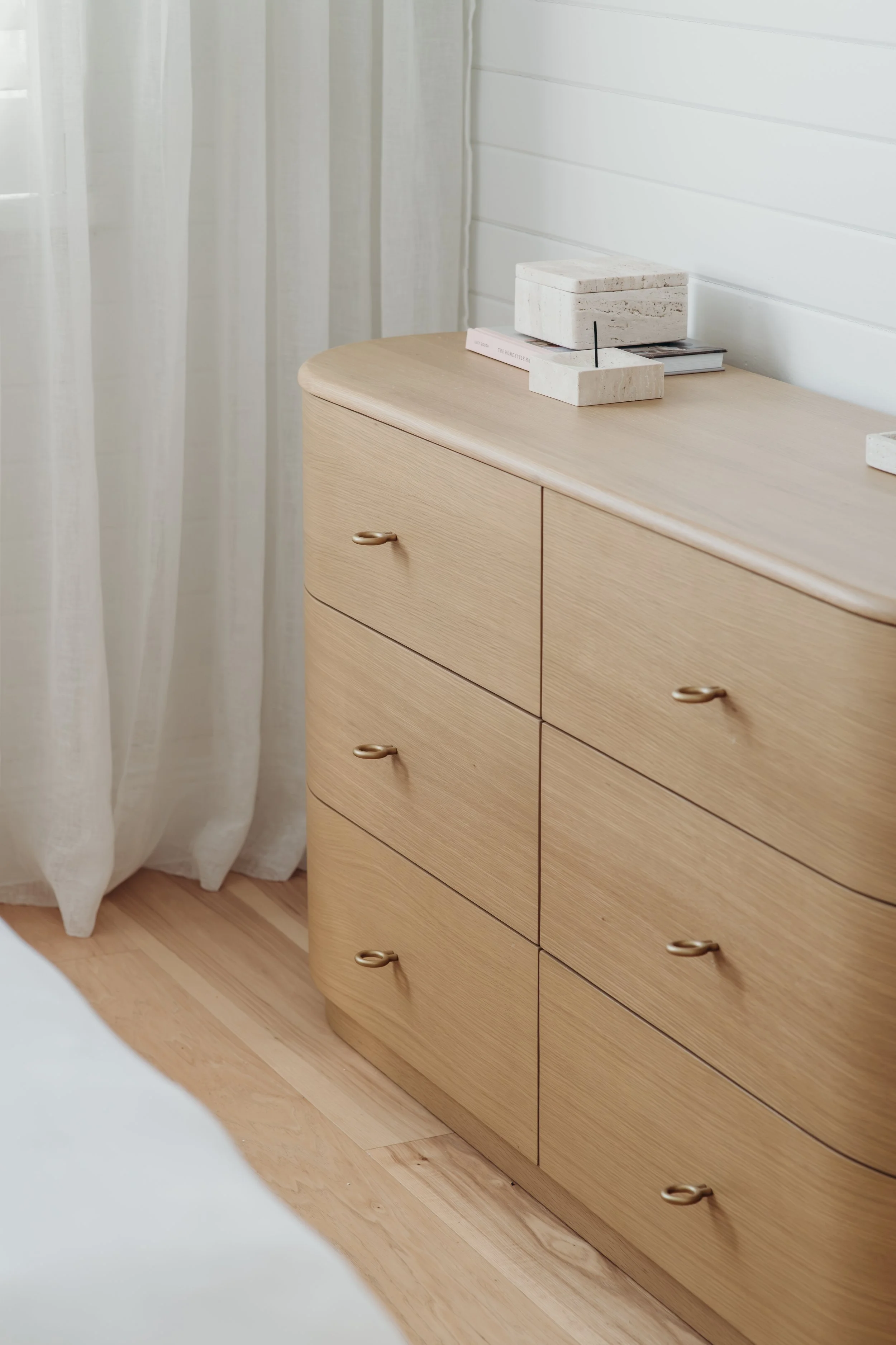 Minimalist light wood dresser with brass ring pulls, stacked books, and a stone box in a bright room with white curtains.